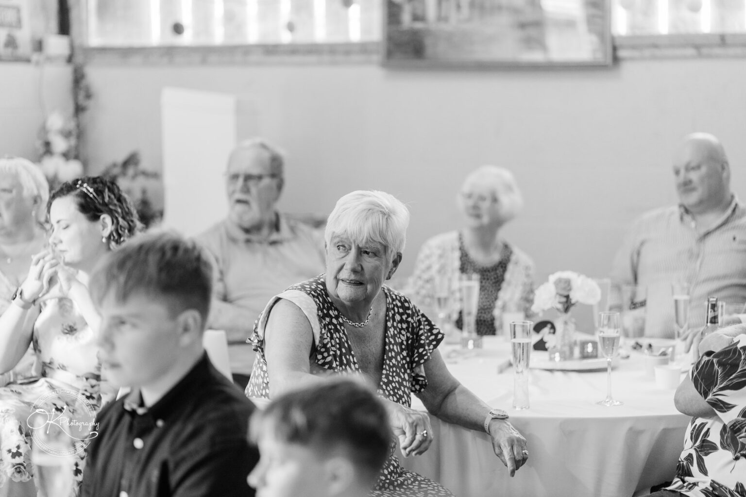 A group of guests seated at tables during a wedding reception, with an older woman in a black and white patterned dress looking thoughtfully.