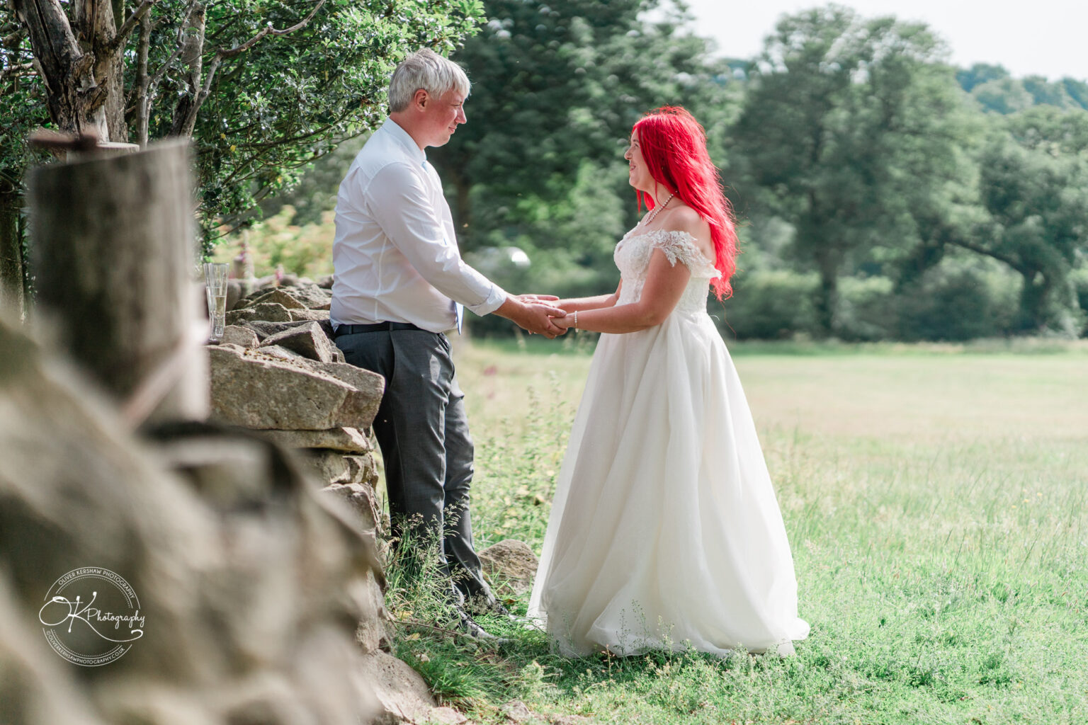 A couple holds hands near a stone wall in an outdoor setting, with a green field and trees in the background. The bride has long red hair and is wearing a white gown.