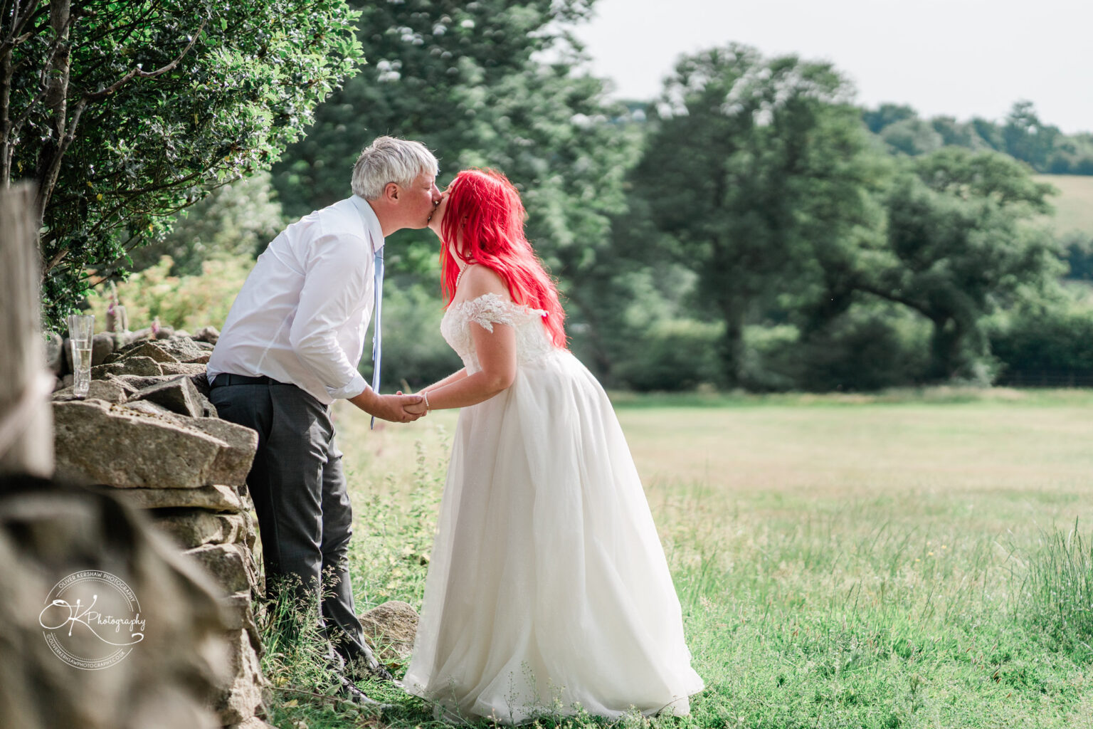 A couple shares a kiss outdoors, surrounded by greenery, with the bride wearing a white gown and the groom in a light grey suit.