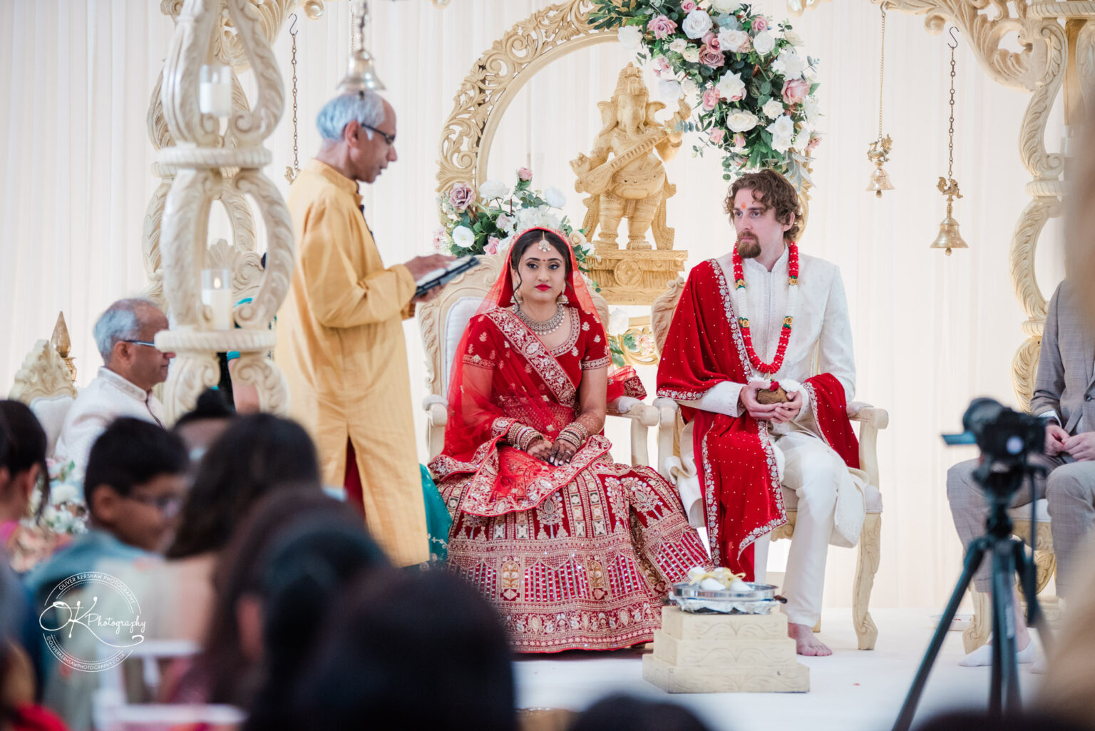 A bride in a red and gold wedding attire sits beside a groom in a white and red outfit, during a ceremony at Winstanley House.