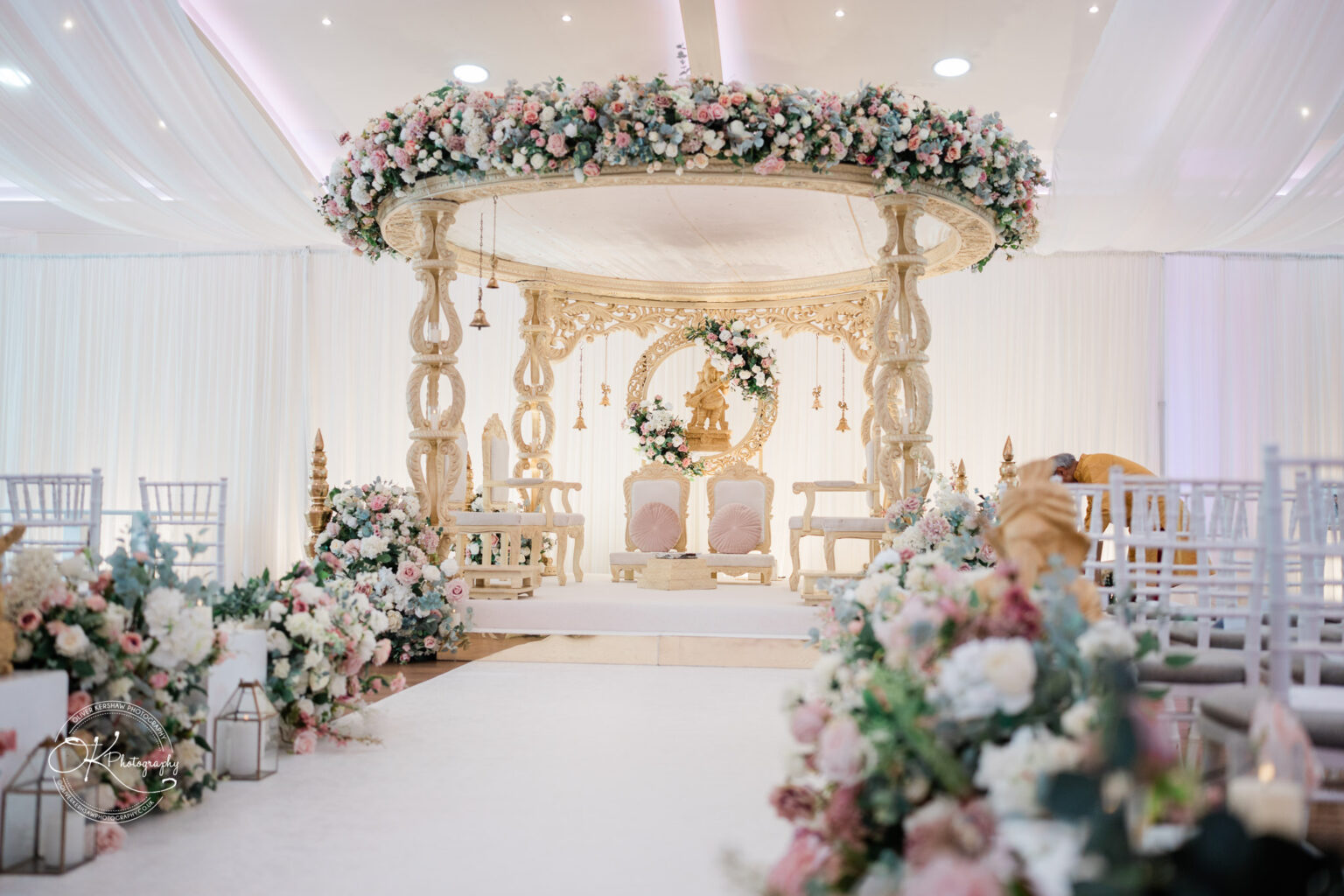 A beautifully decorated wedding venue featuring an elegant floral arch, a decorative seating area for the couple, and floral arrangements lining the aisle.