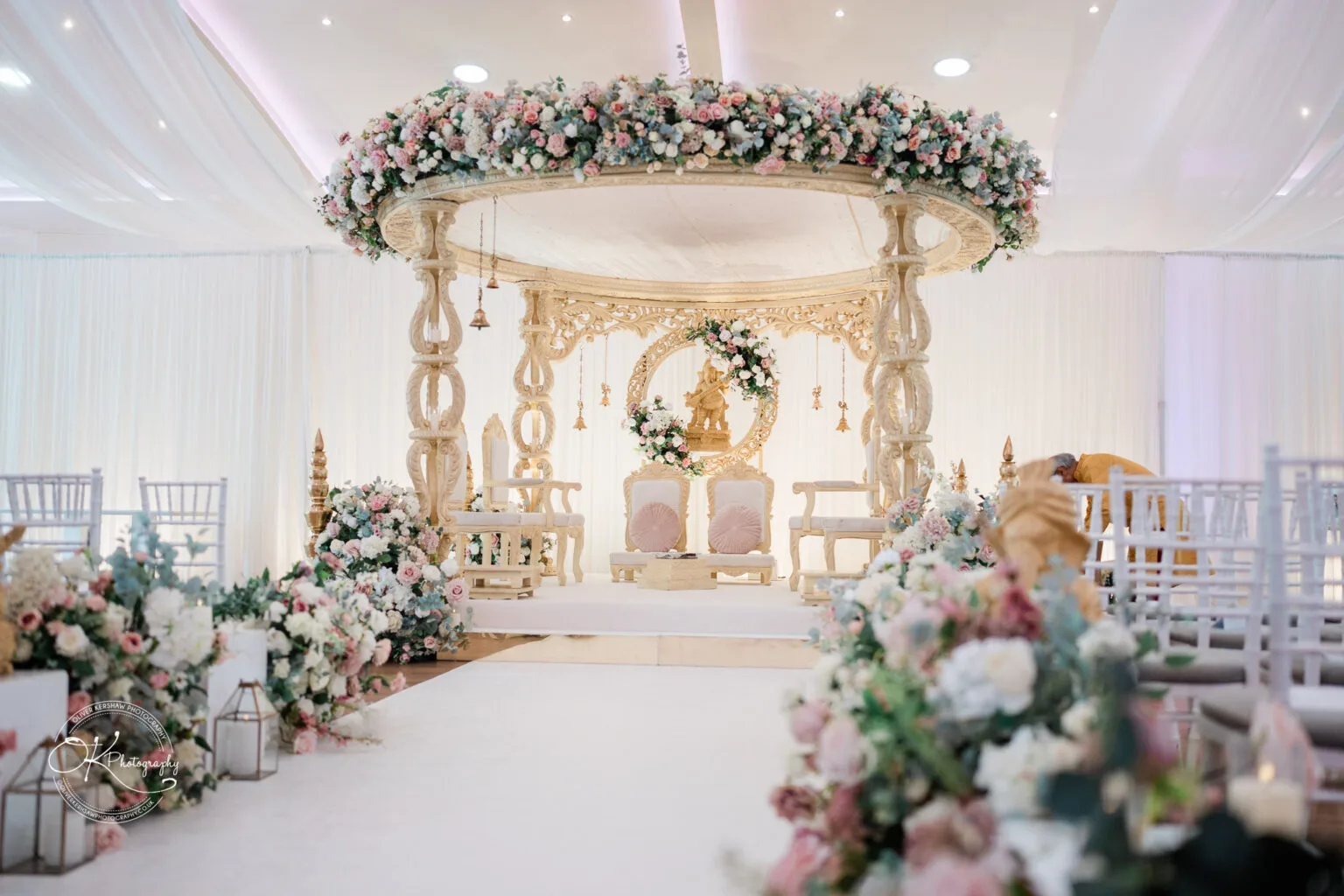 Roshni & Joseph-wedding -Winstanley House-Photography By Oliver Kershaw - OK501739 A beautifully decorated wedding venue featuring an elegant floral arch, a decorative seating area for the couple, and floral arrangements lining the aisle.