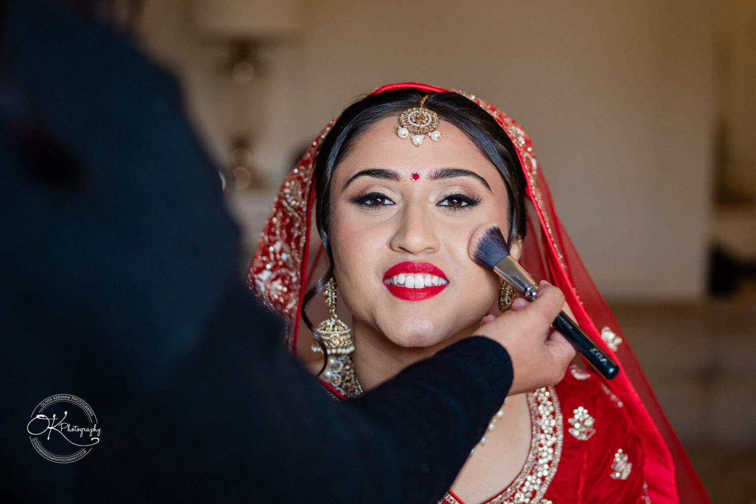 A bride in traditional attire, wearing a red veil and jewelry, has her makeup applied, smiling at the camera.