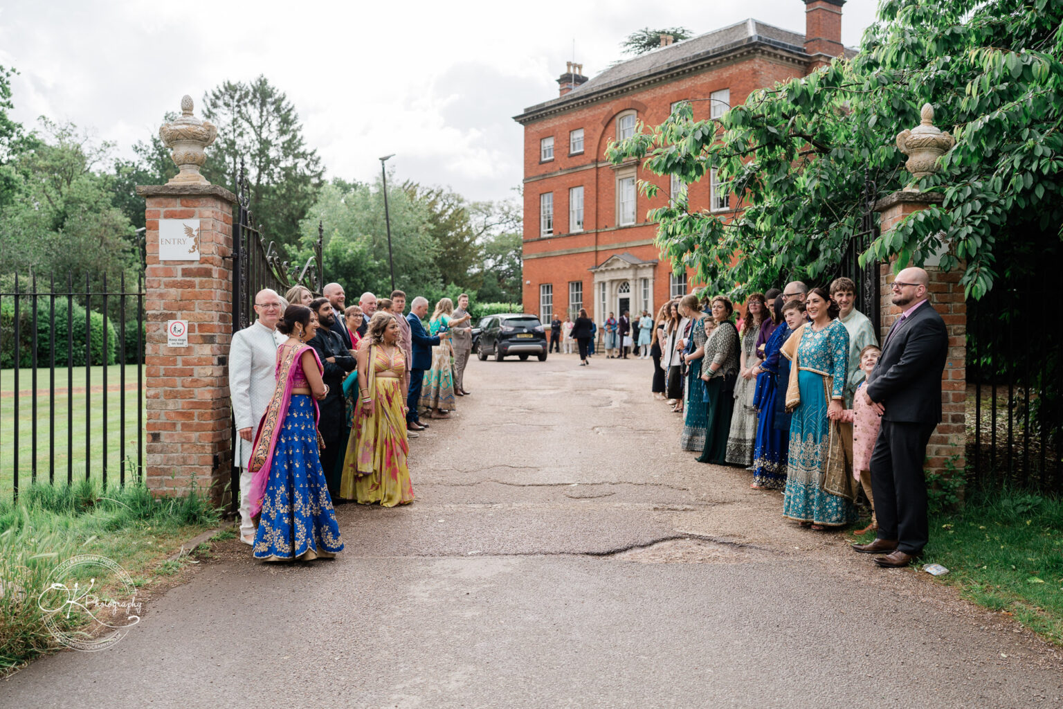 Guests dressed in vibrant traditional attire are gathered along a driveway outside Winstanley House, welcoming an arriving wedding party.