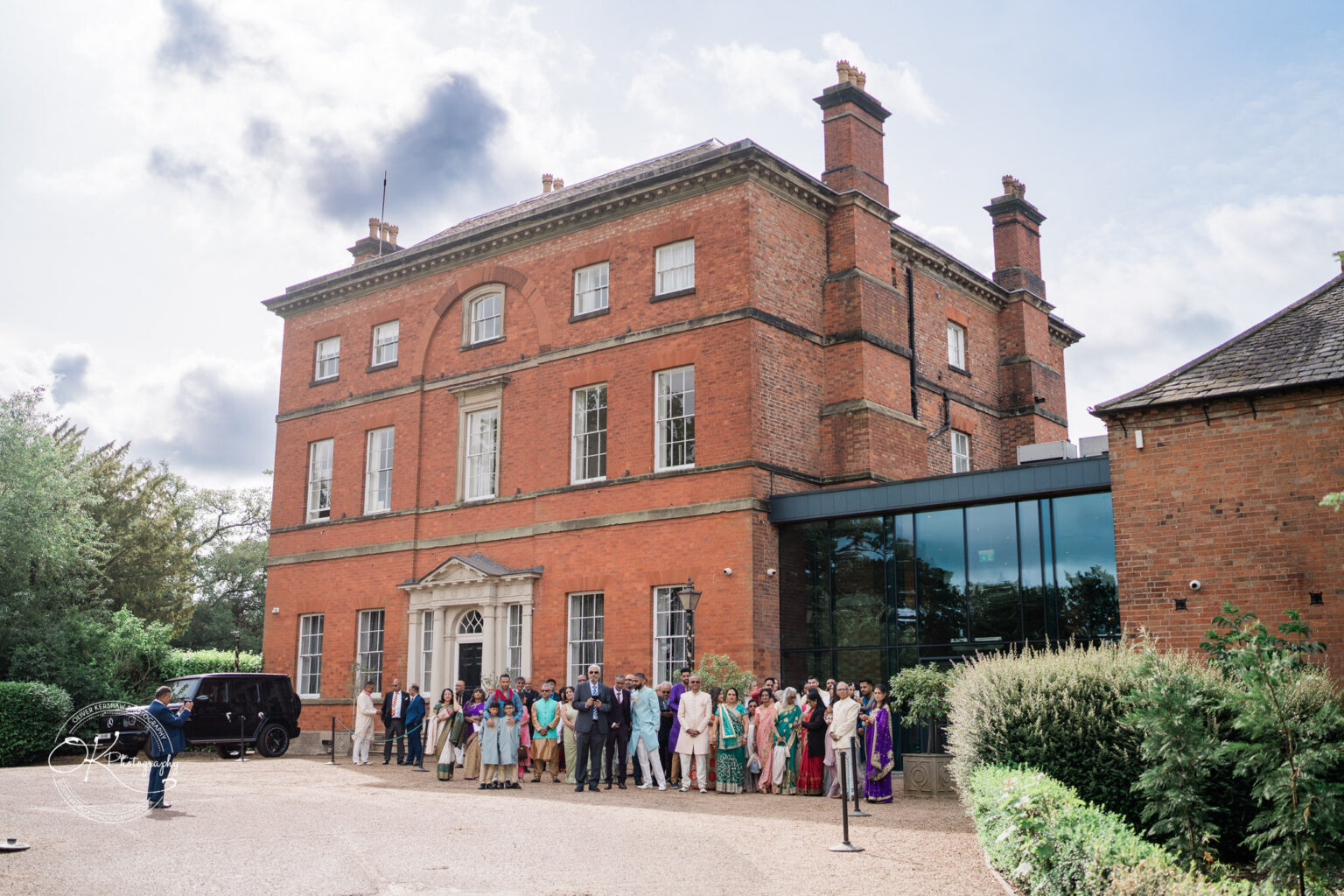 A large group of people gathered outside Winstanley House, showcasing a mix of formal and traditional attire in front of the impressive red brick building.