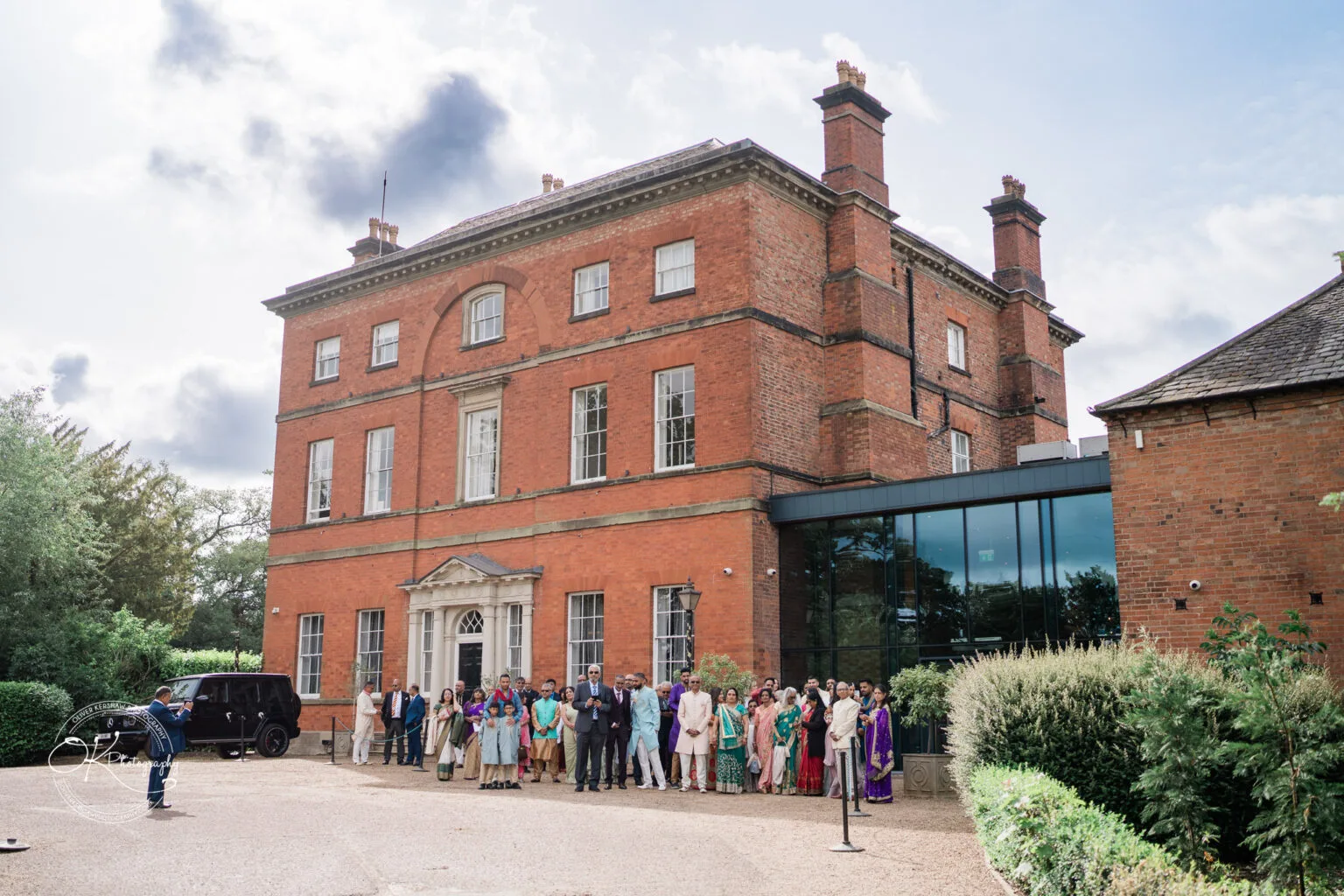 Roshni & Joseph-wedding -Winstanley House-Photography By Oliver Kershaw - OK501869 A large group of people gathered outside Winstanley House, showcasing a mix of formal and traditional attire in front of the impressive red brick building.