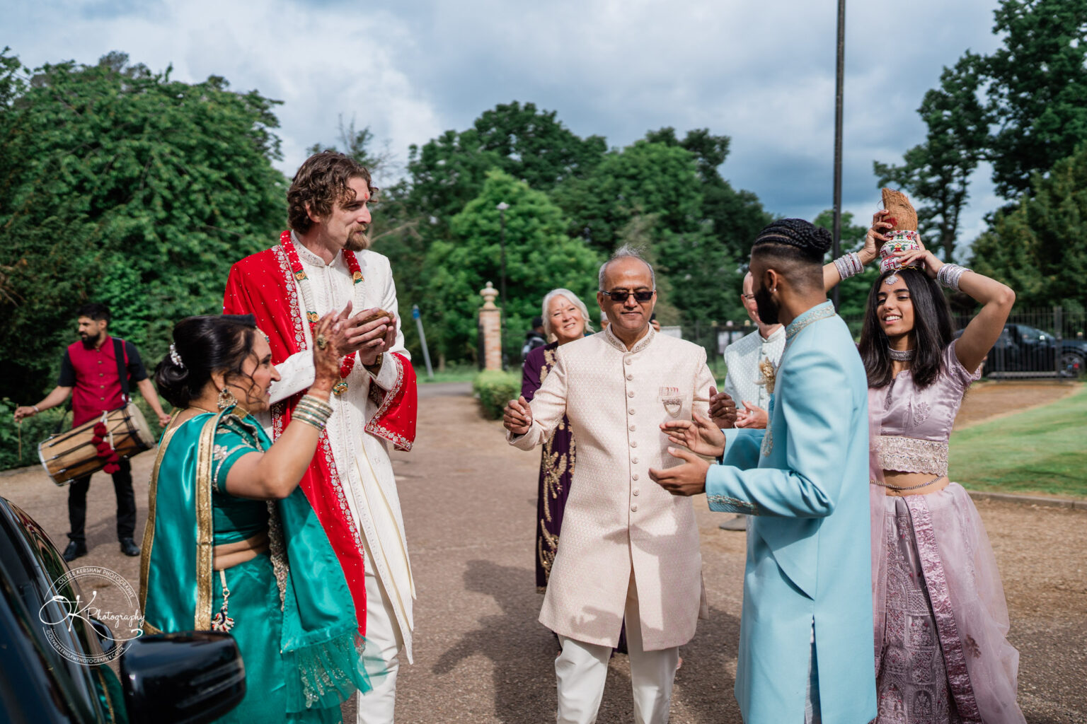 A diverse group of wedding guests celebrate outside Winstanley House, dressed in traditional attire, with joyful expressions and festive gestures.
