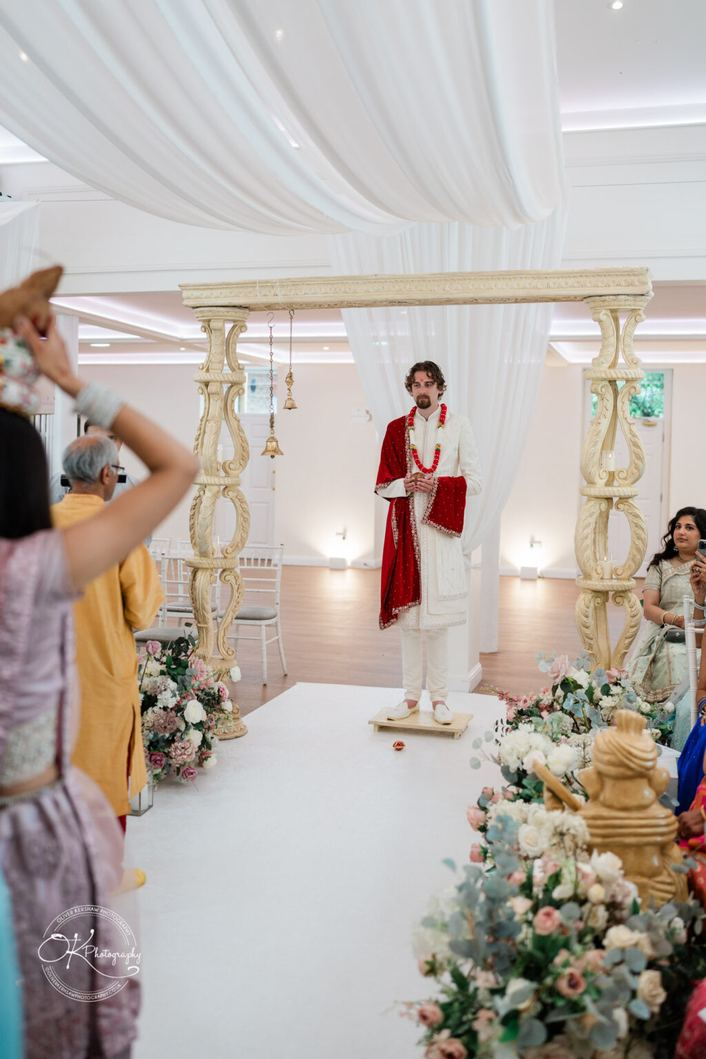 A groom in traditional attire stands under an ornate floral wedding arch, surrounded by guests in colourful garments during a ceremony.