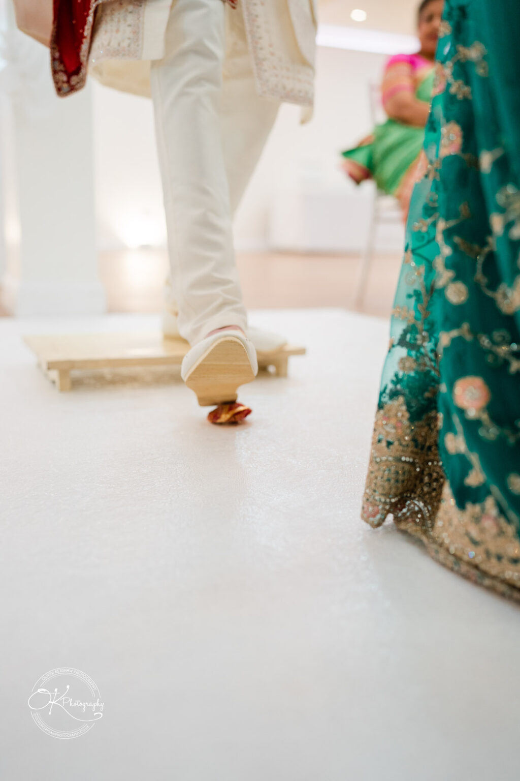 A groom steps onto a floral-adorned board, with a decorated bridal party in vibrant attire visible in the background.