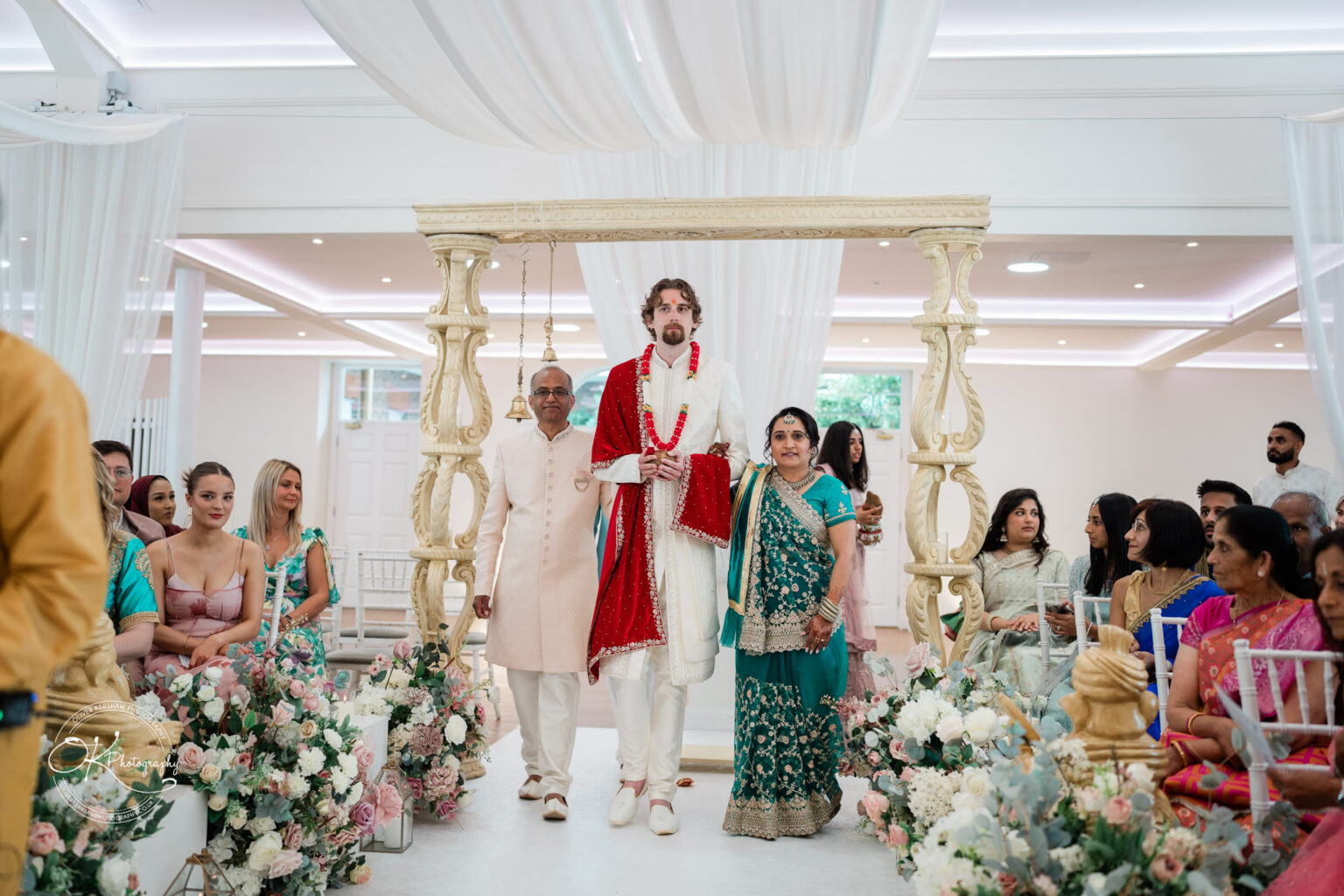 A groom in traditional attire stands at a beautifully decorated wedding altar, flanked by his parents, amidst floral arrangements and guests.