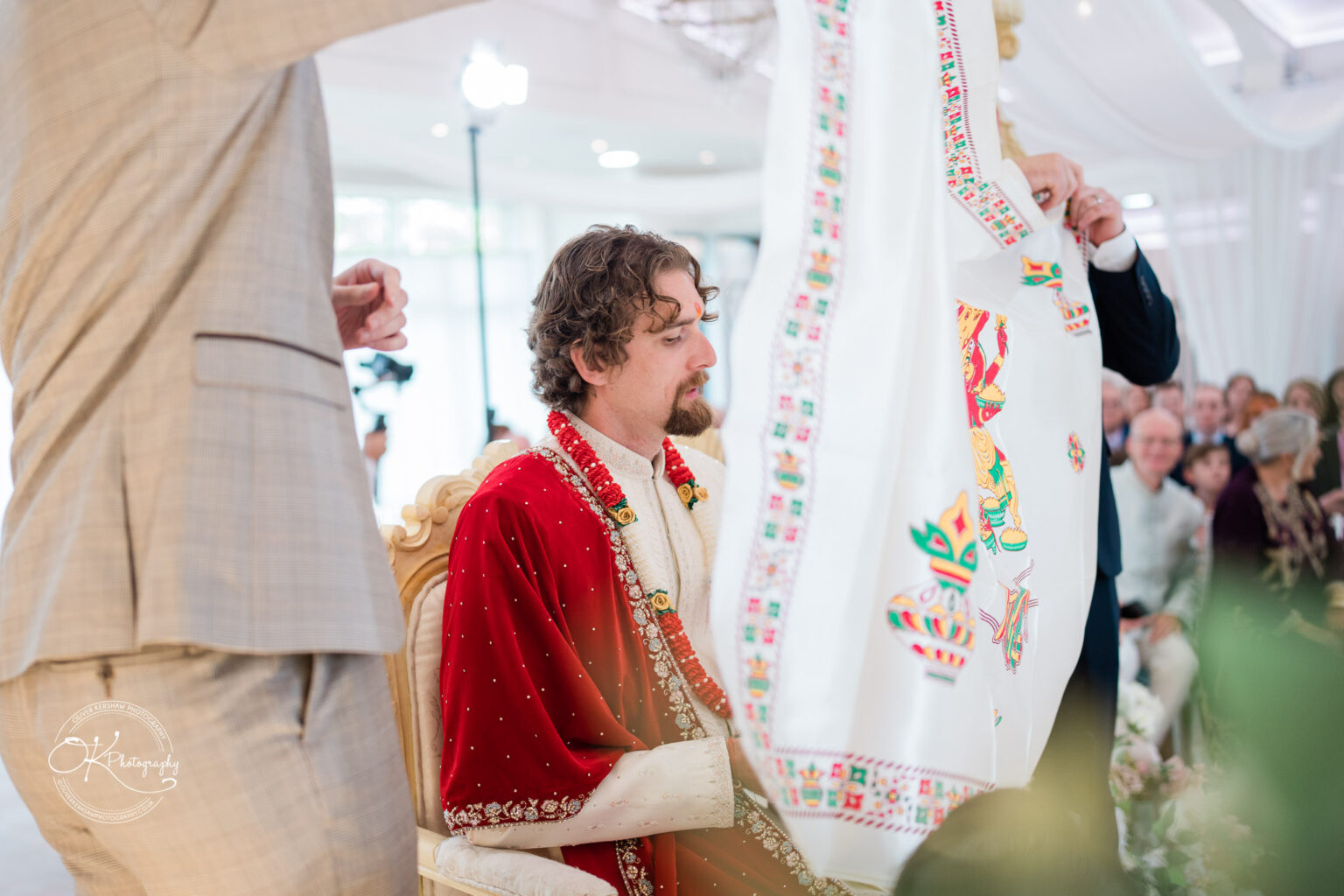 A groom in traditional attire sits at a decorated altar during a wedding ceremony, with guests watching in the background.