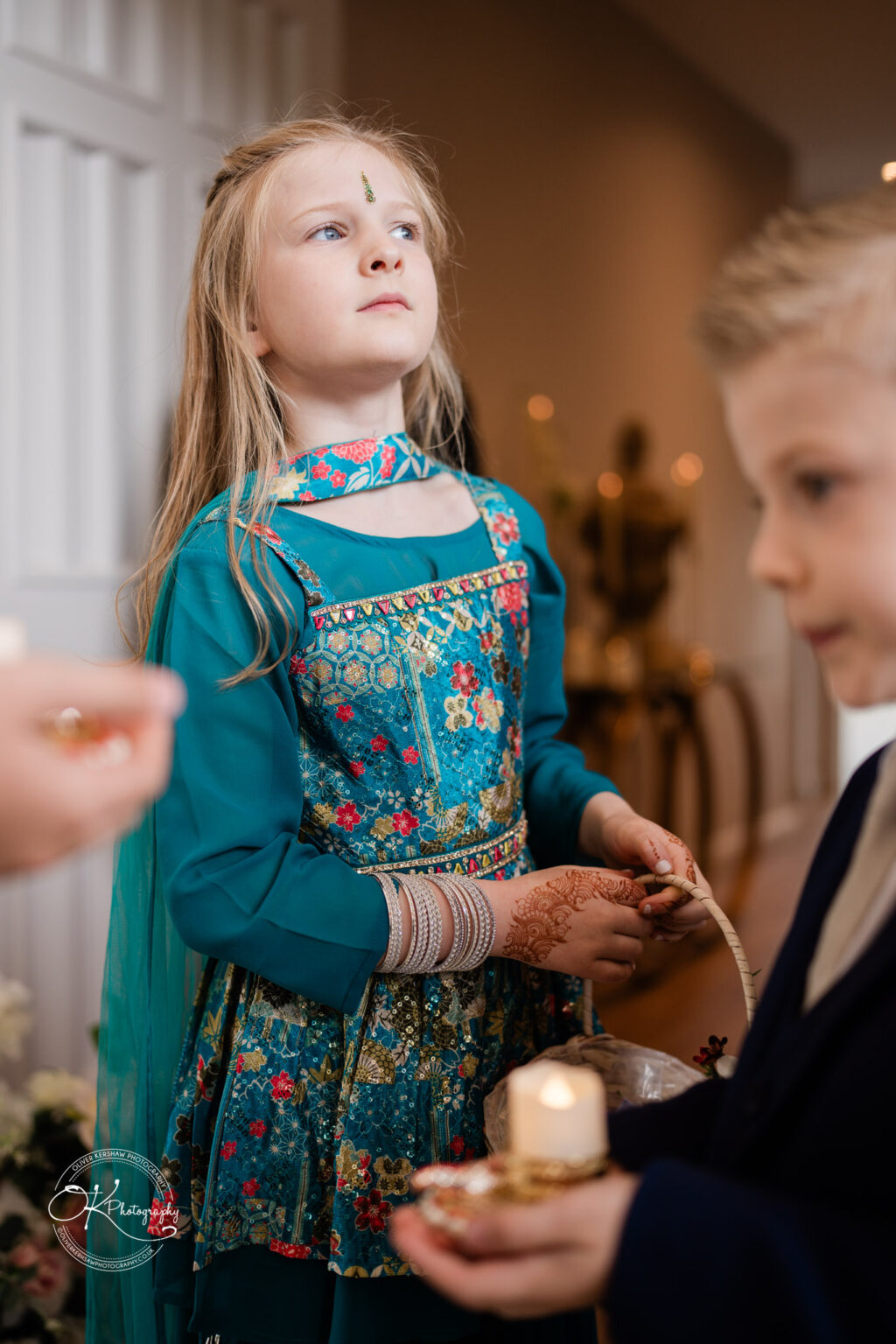 A young girl in traditional attire holds a basket and looks thoughtfully, with a boy nearby in formal wear.