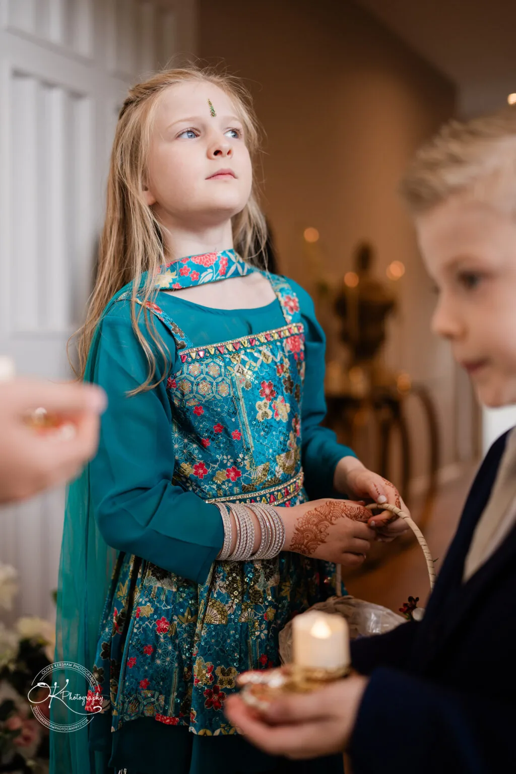Roshni & Joseph-wedding -Winstanley House-Photography By Oliver Kershaw - OK502241 A young girl in traditional attire holds a basket and looks thoughtfully, with a boy nearby in formal wear.