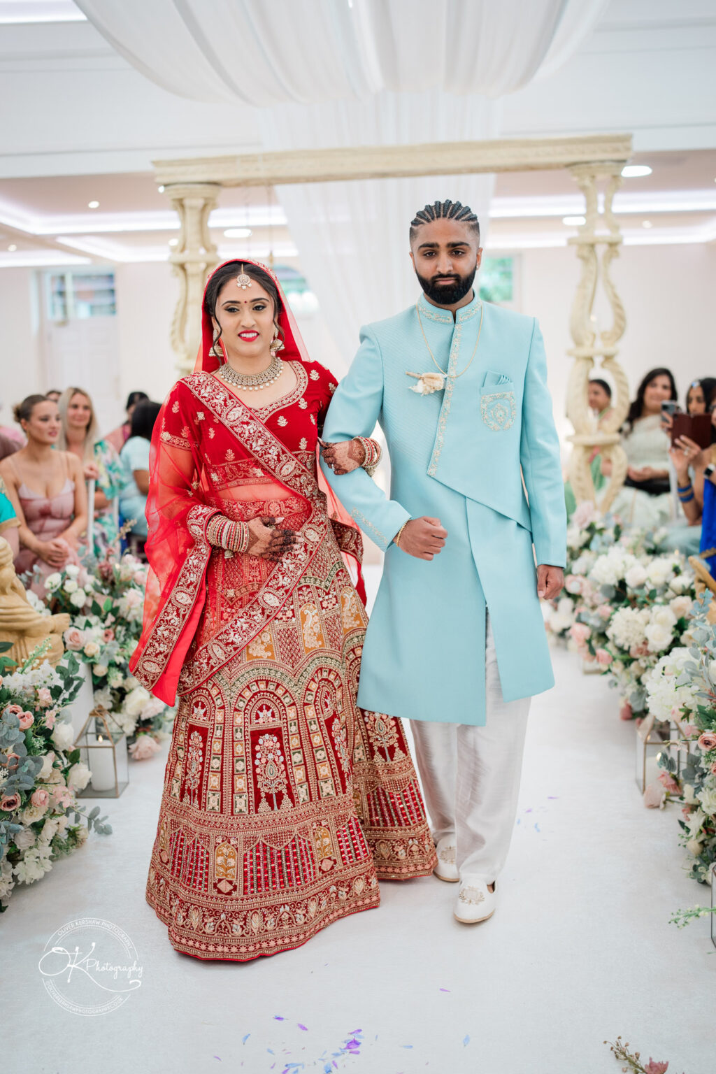 A bride in a red and gold traditional outfit and a groom in a light blue sherwani walk down the aisle, surrounded by floral decorations.