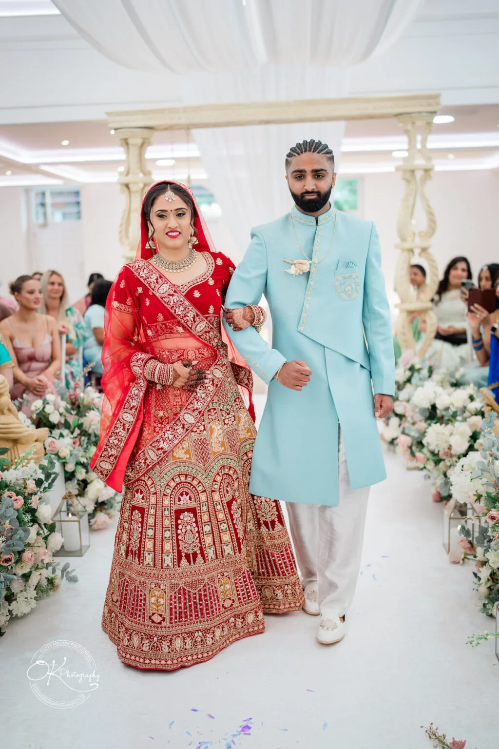 Roshni & Joseph-wedding -Winstanley House-Photography By Oliver Kershaw - OK502297 A bride in a red and gold traditional outfit and a groom in a light blue sherwani walk down the aisle, surrounded by floral decorations.