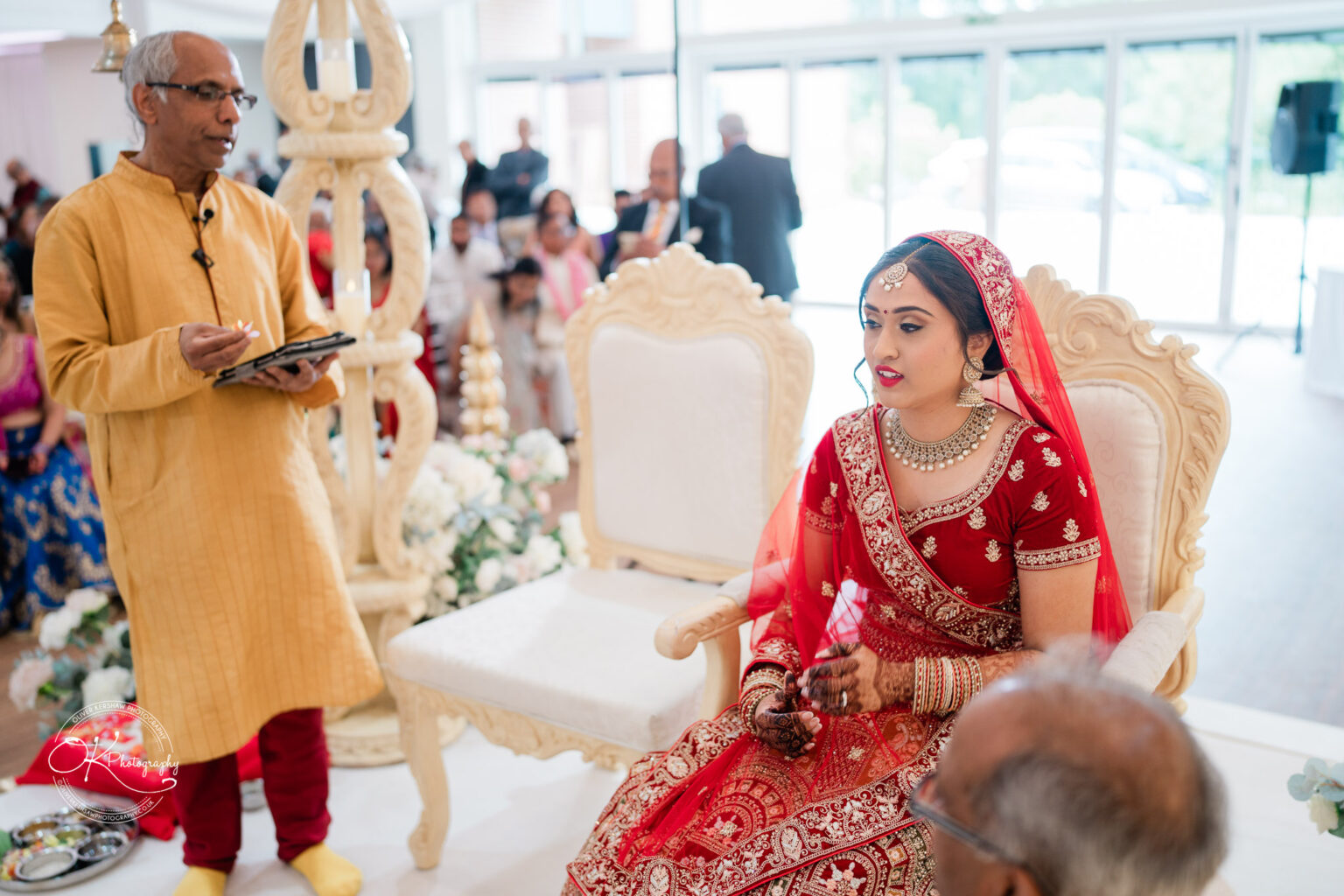 A bride in traditional red attire adorned with jewellery sits in a decorative chair, while a man in a yellow outfit speaks, surrounded by wedding guests.