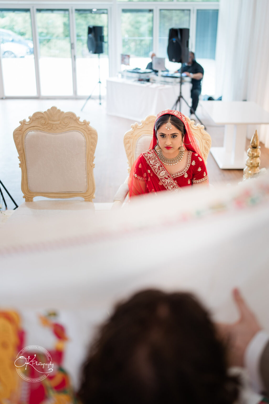 A bride in a red and gold outfit sits before a decorated altar, with a veil draped in front, in a bright wedding venue.