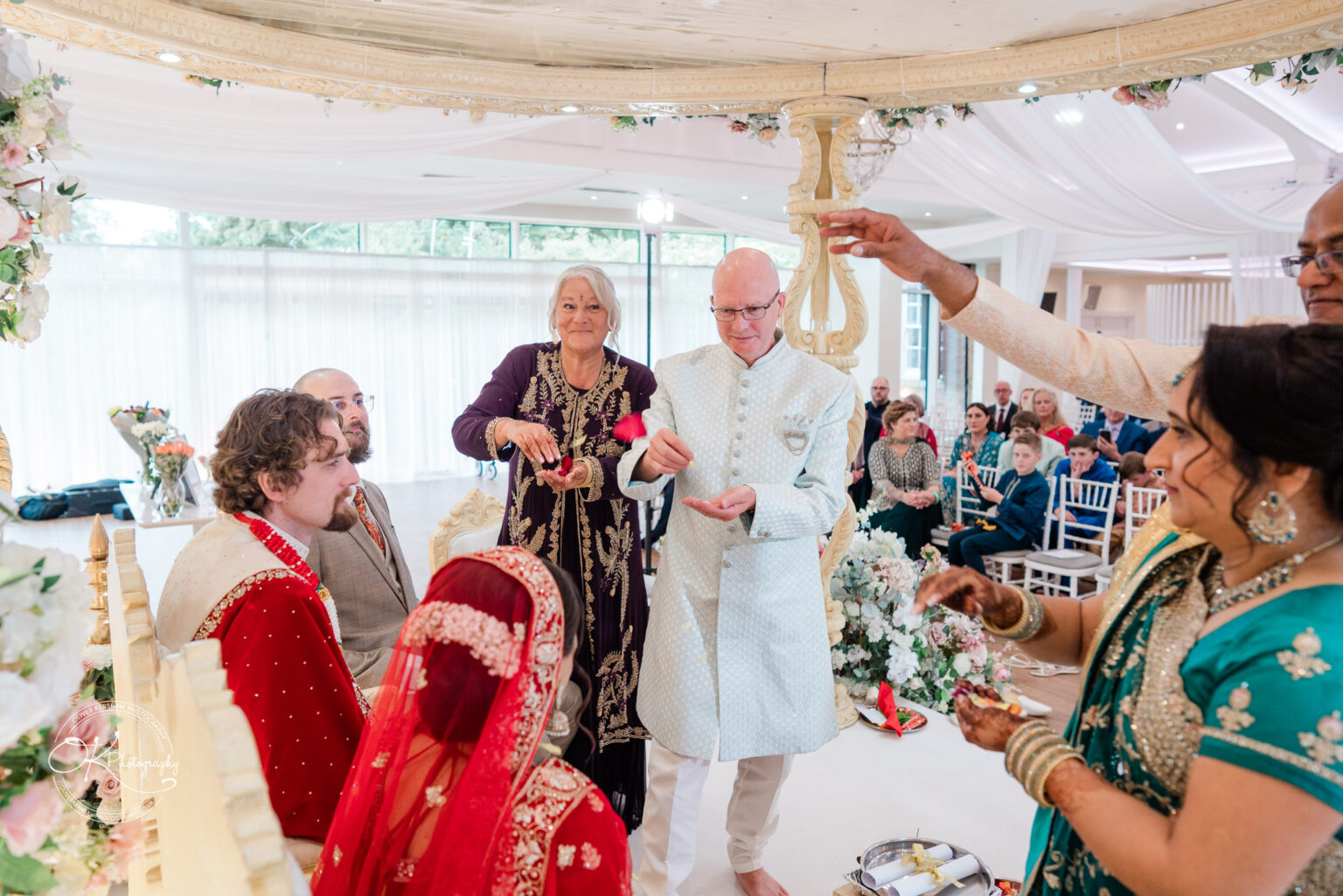 A wedding ceremony at Winstanley House, featuring a bride in red and gold attire, a groom, and family members participating in traditional rituals.