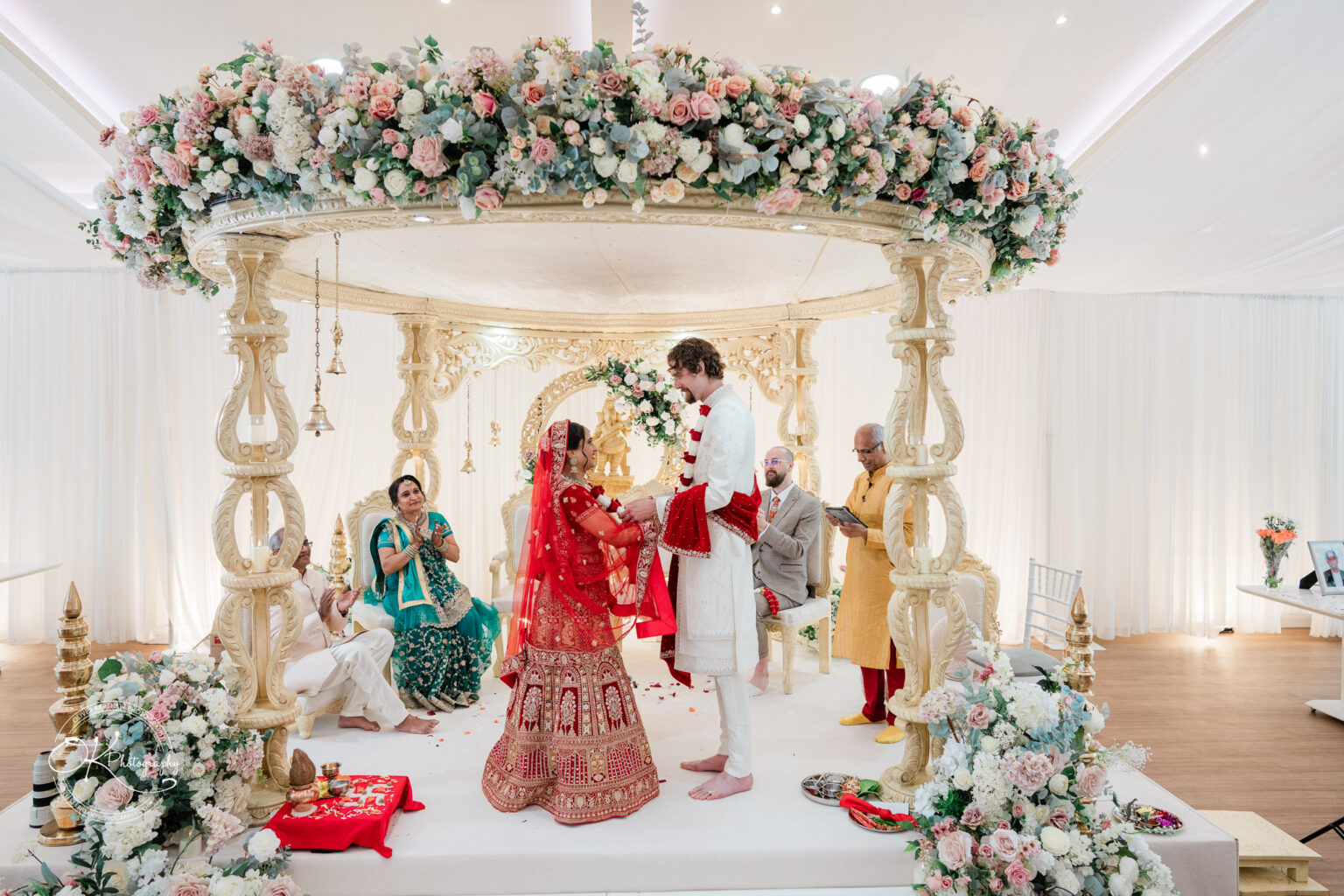 A bride in a red traditional outfit and groom in white attire stand under a floral wedding canopy, surrounded by guests in festive clothing.
