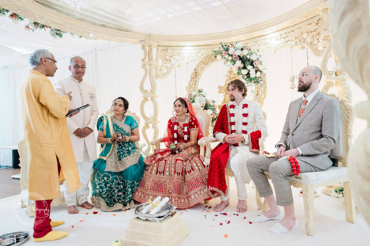 A bride and groom seated in traditional attire during a wedding ceremony, surrounded by family and a officiant.