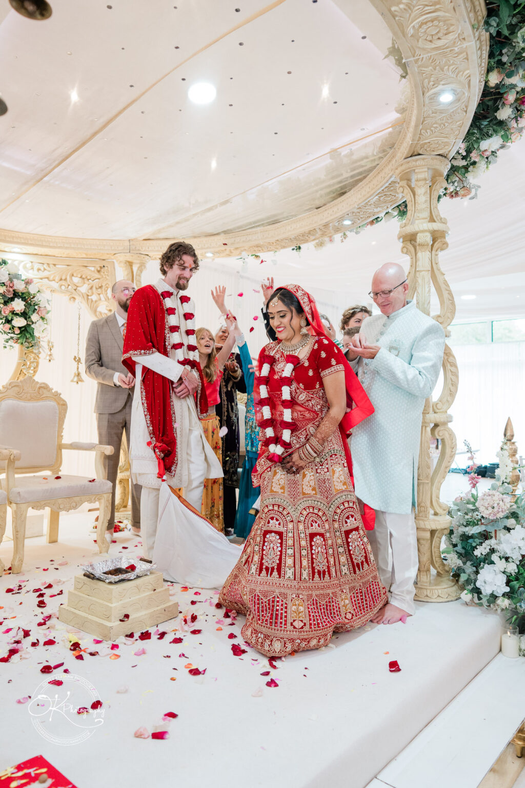 A couple in traditional wedding attire stands under an ornate canopy, surrounded by guests, with rose petals scattered on the floor.