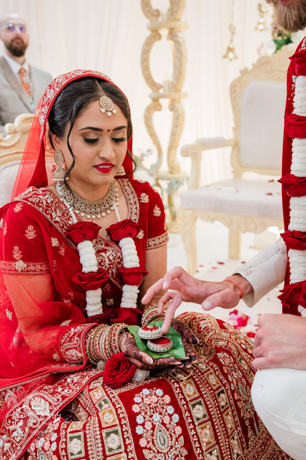 A bride in a vibrant red wedding outfit seated during a ceremony, adorned with jewelry and floral decorations, receives a ring.