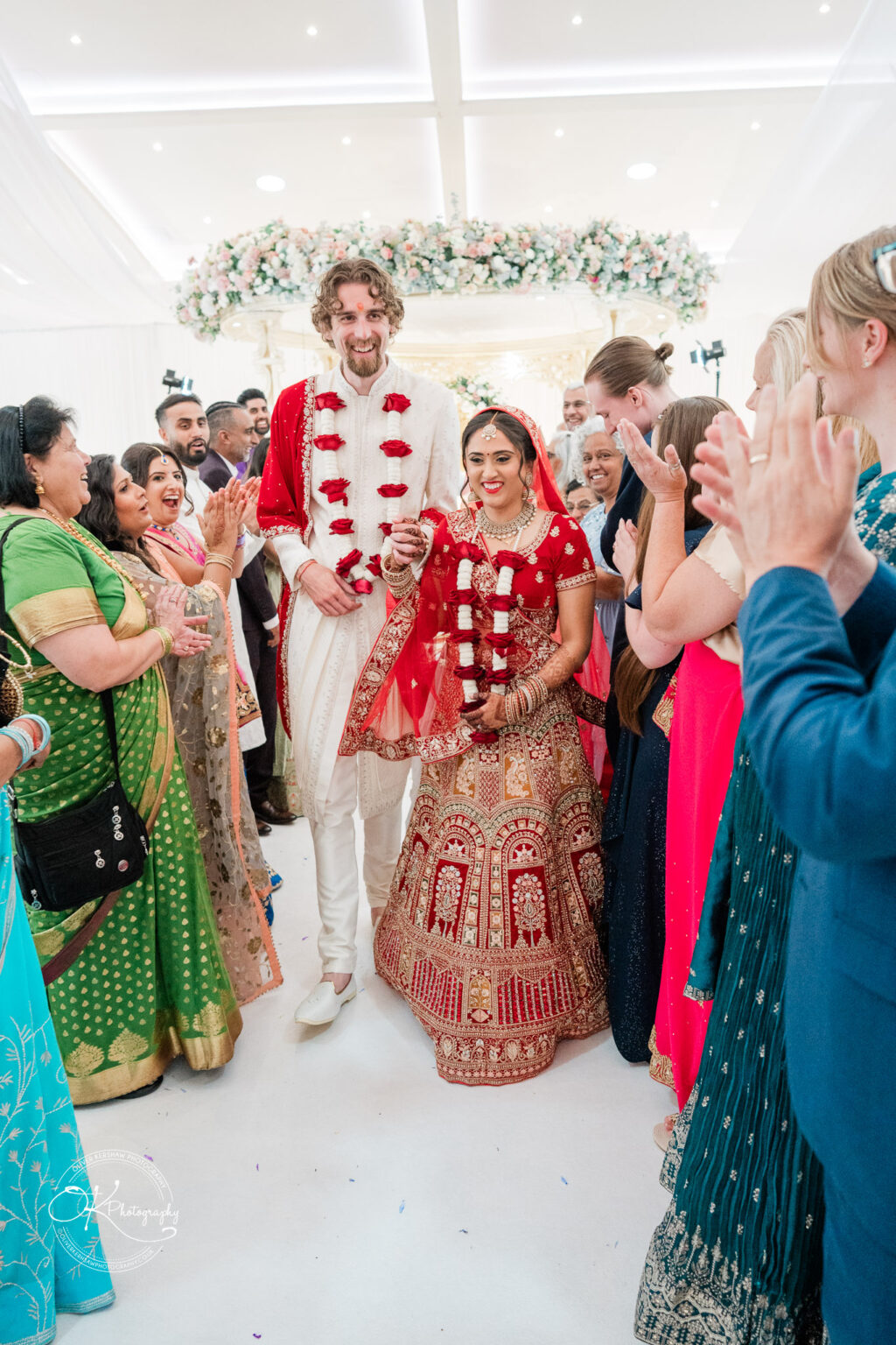 A groom in a white sherwani and a bride in a red and gold wedding outfit, both adorned with flower garlands, walk together amid a joyous crowd.