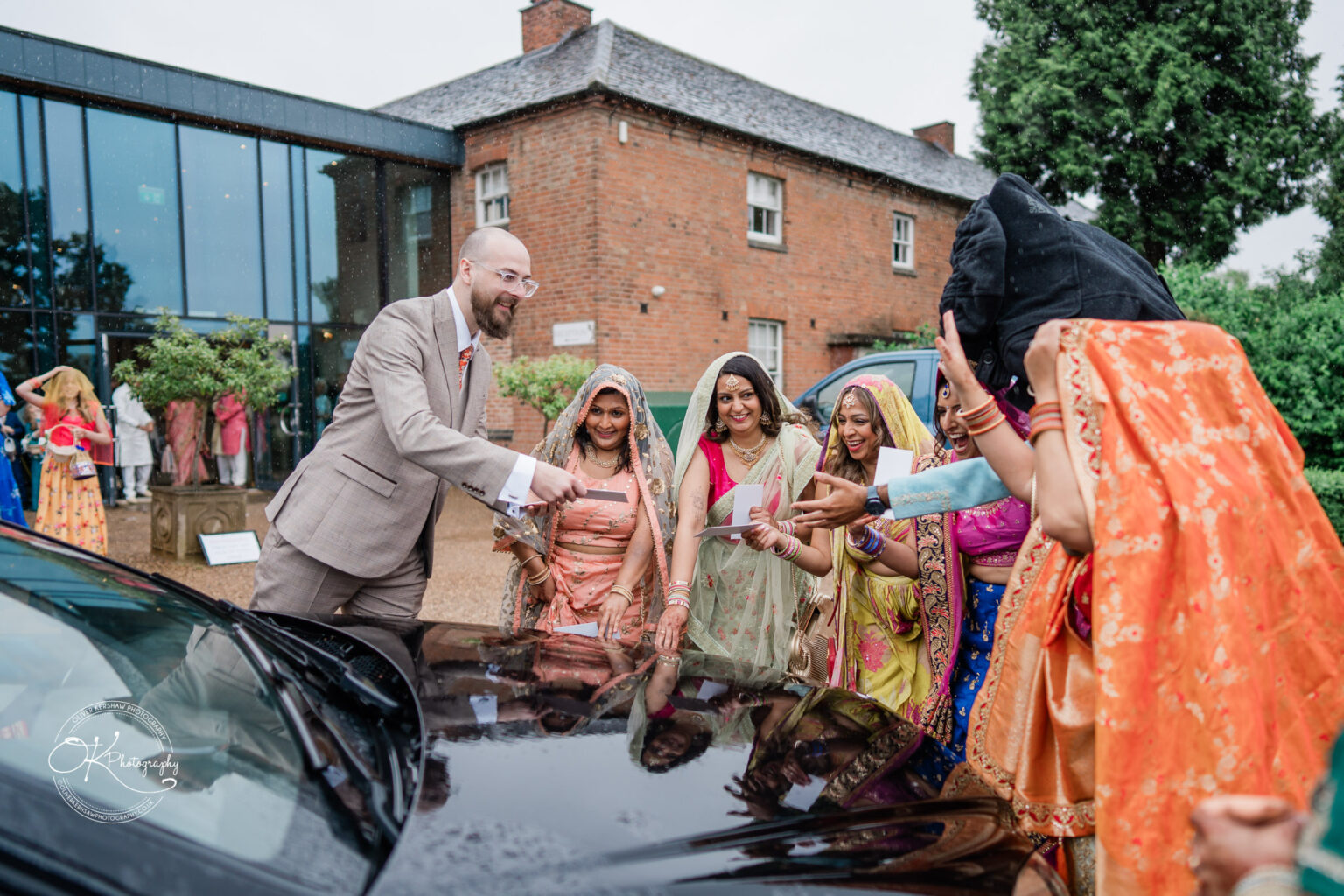 A groom greets bridesmaids in colorful traditional attire outside Winstanley House, with a rainy background and elegant venue.