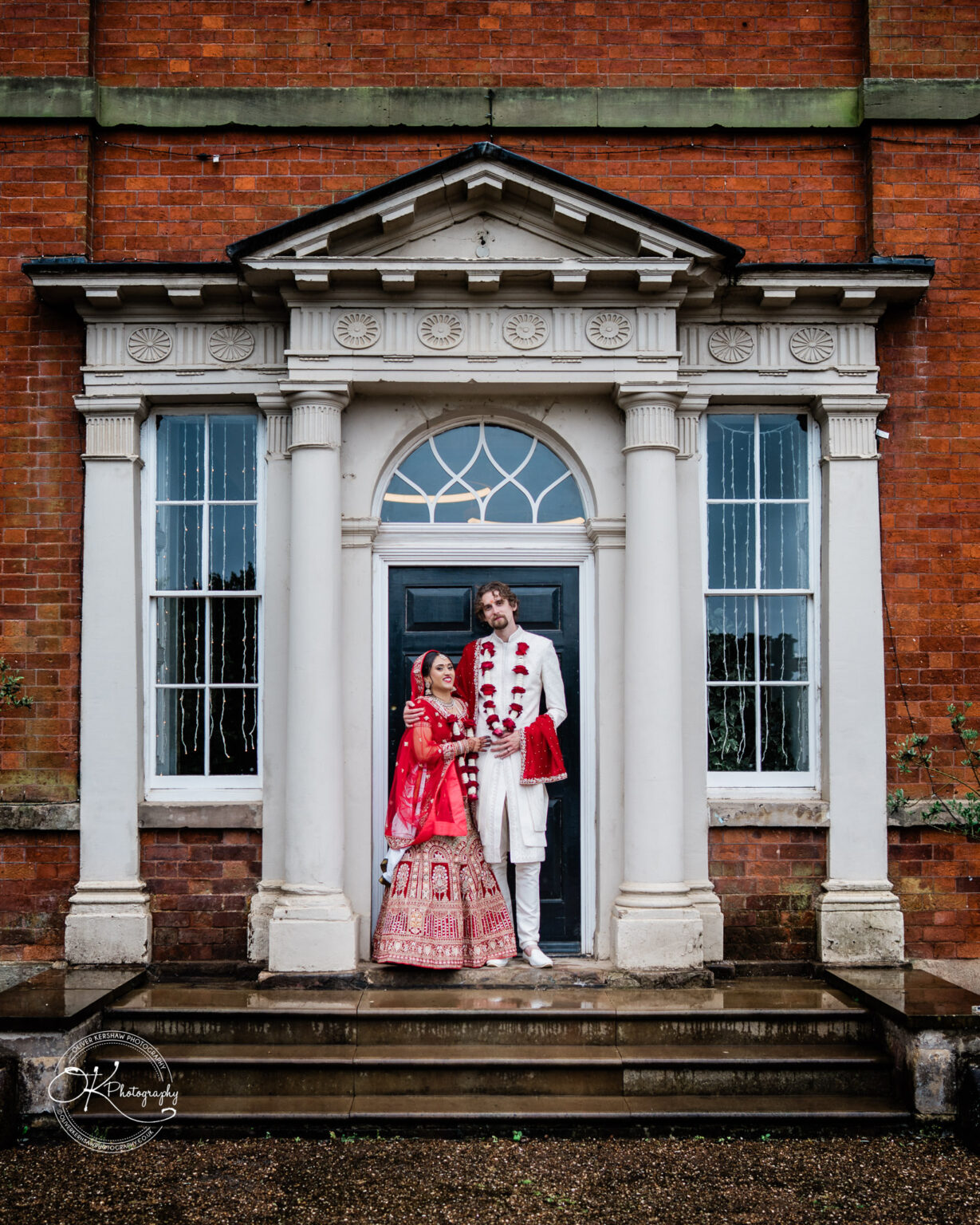 A bride in a red and gold wedding outfit and a groom in traditional attire stand together on the steps of Winstanley House.