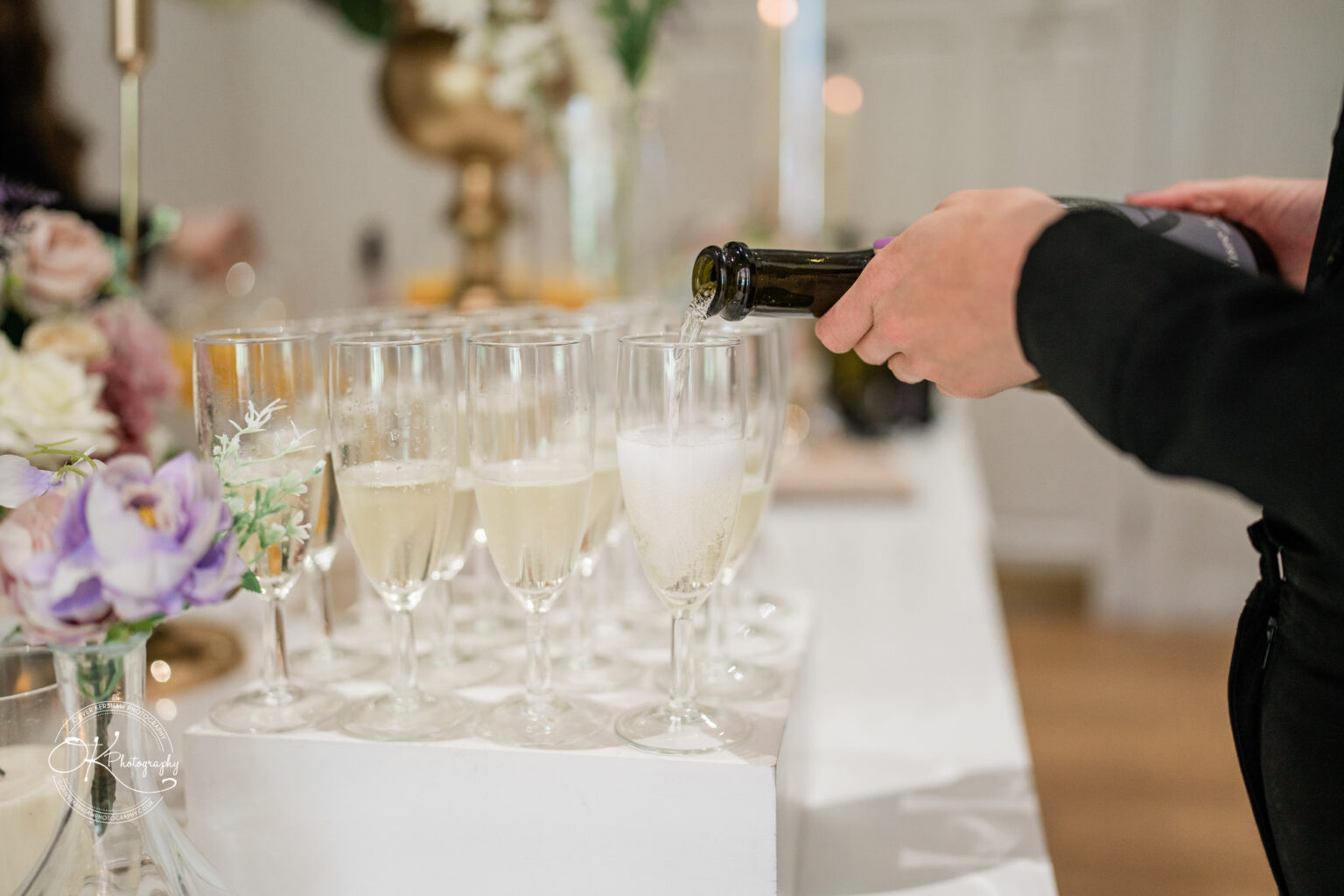 A hand pours champagne into a glass, with several filled glasses on a white table, surrounded by floral arrangements.