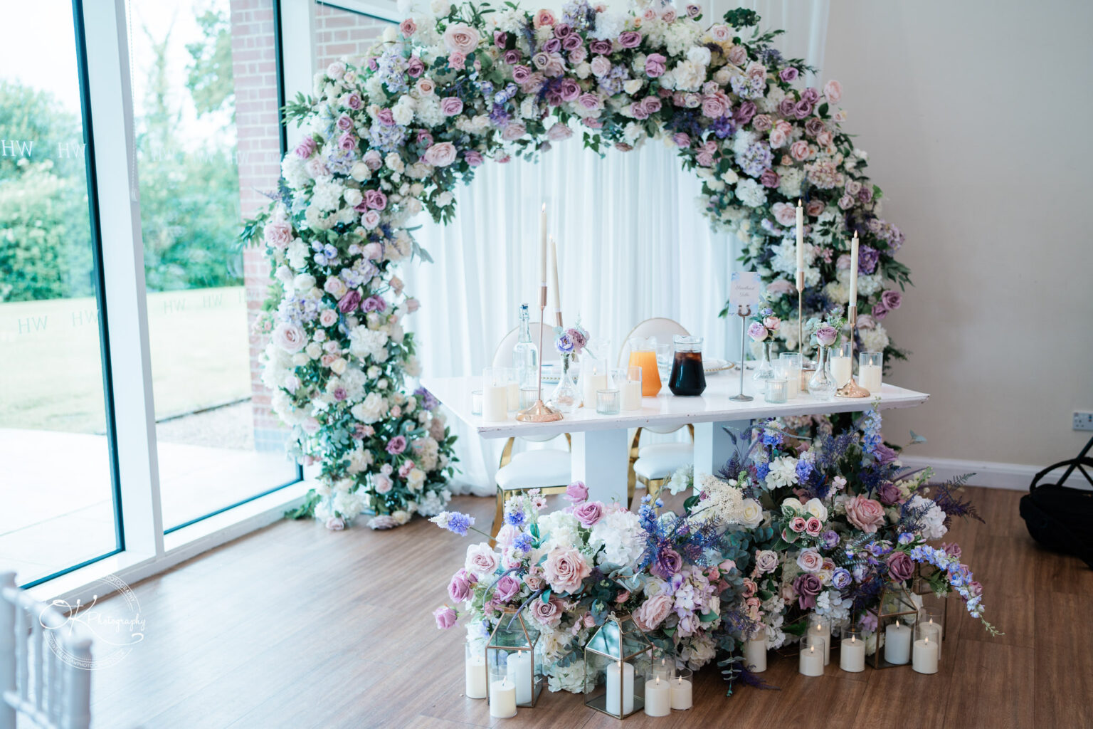 A beautifully decorated wedding reception area featuring a floral arch with purple and white flowers, a table set with candles and soft floral arrangements.