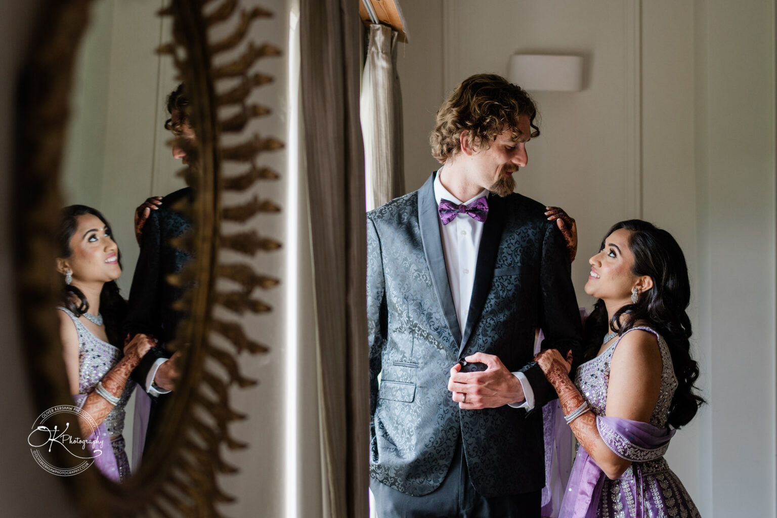 A bride in a traditional outfit smiles at her groom in a formal suit, both standing near a mirror in Winstanley House.