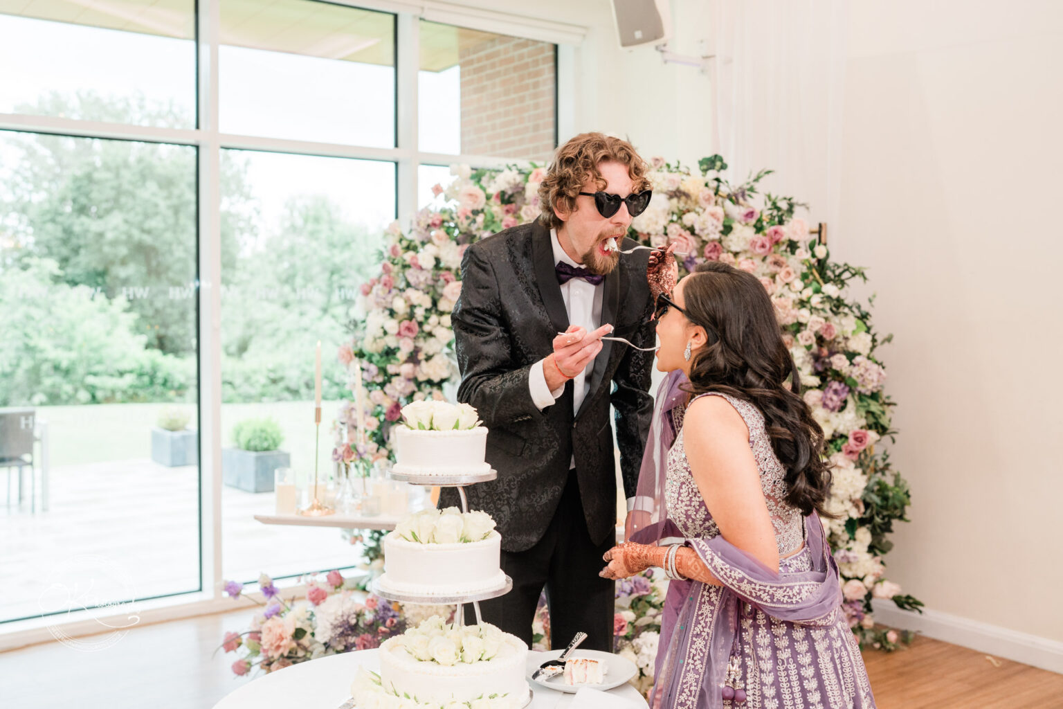 A couple playfully feeding each other wedding cake, surrounded by a floral backdrop, in a bright venue.