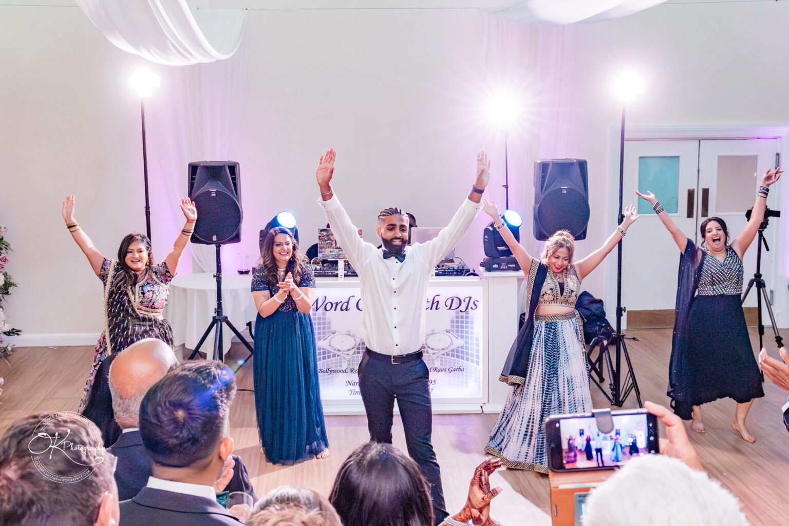 A joyful wedding scene at Winstanley House, featuring a man with raised arms performing with four women dressed in elegant outfits, as guests celebrate.