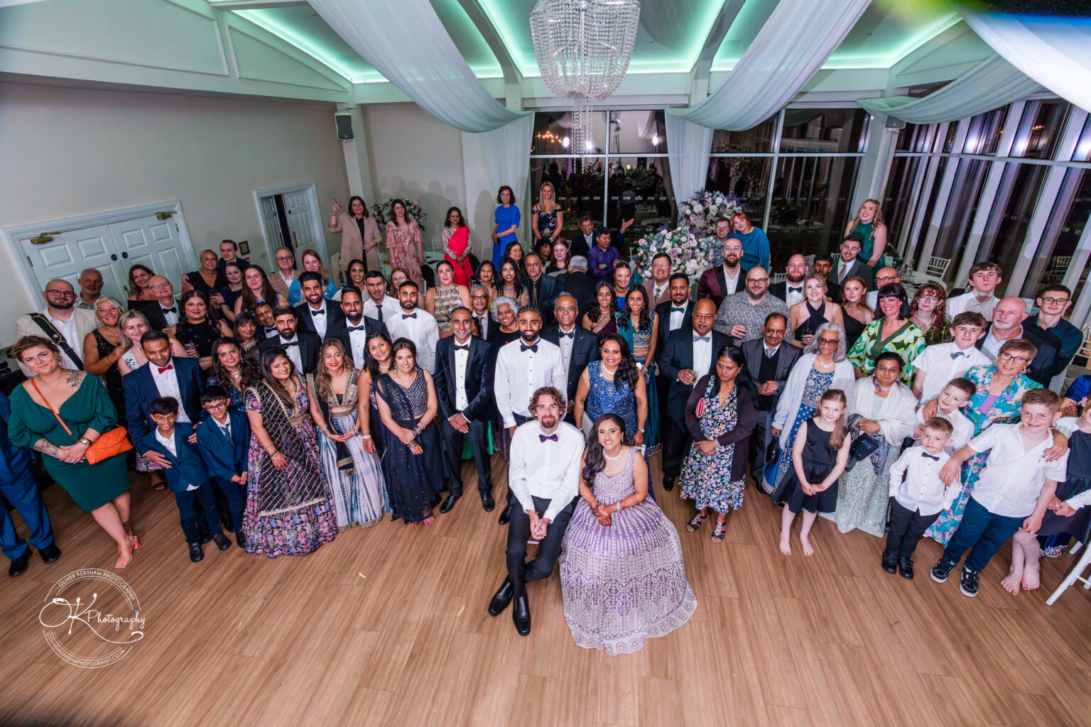 A large group of wedding guests poses for a photo at Winstanley House, with the couple seated in the front among the attendees.