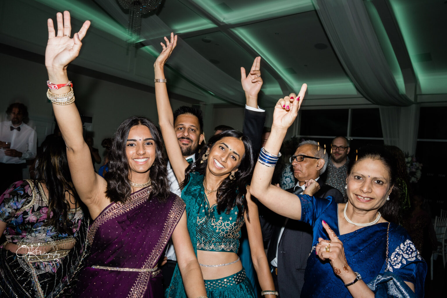 A lively group of guests at a wedding celebration, dressed in vibrant attire, joyfully raising their arms and smiling.