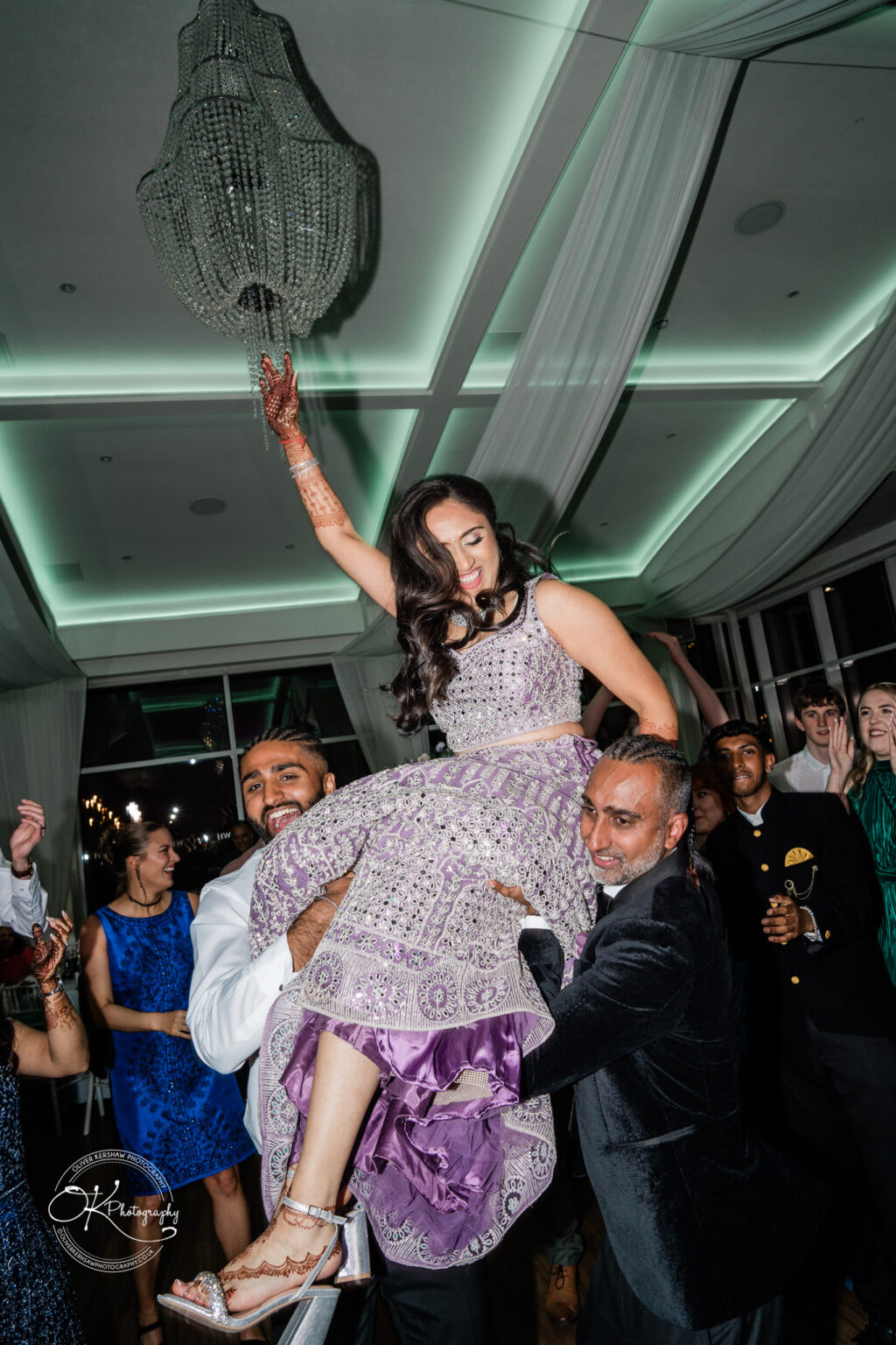 A bride is being lifted in celebration by two men during a wedding reception at Winstanley House, surrounded by guests.