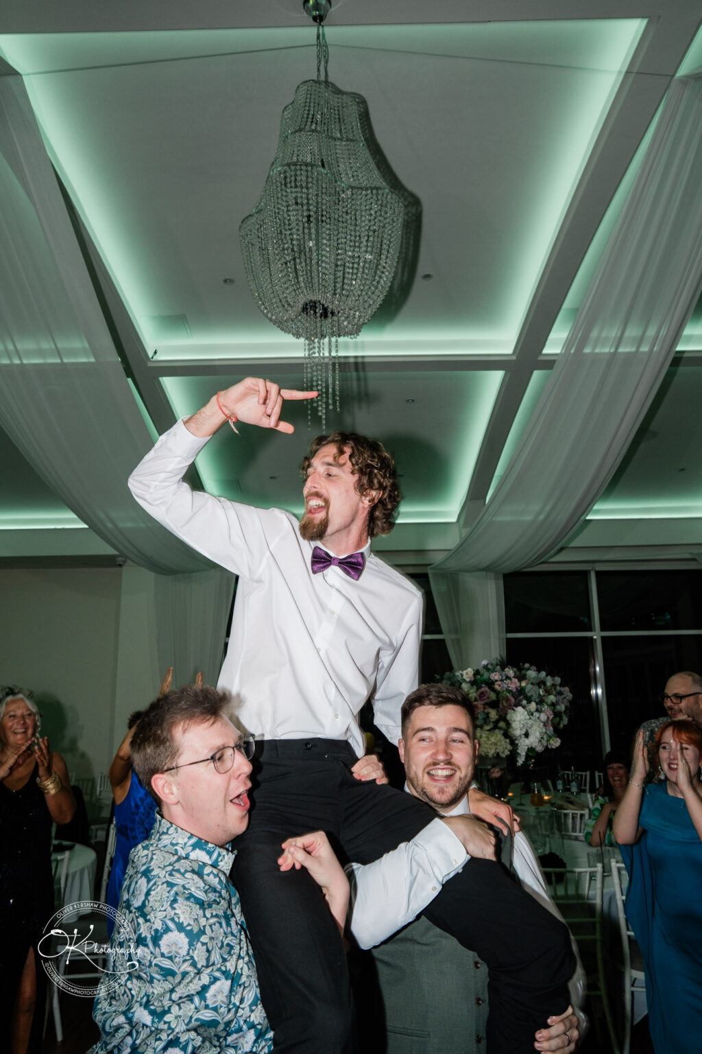 A groom is being held up by two friends during a wedding celebration at Winstanley House, with guests cheering in the background.