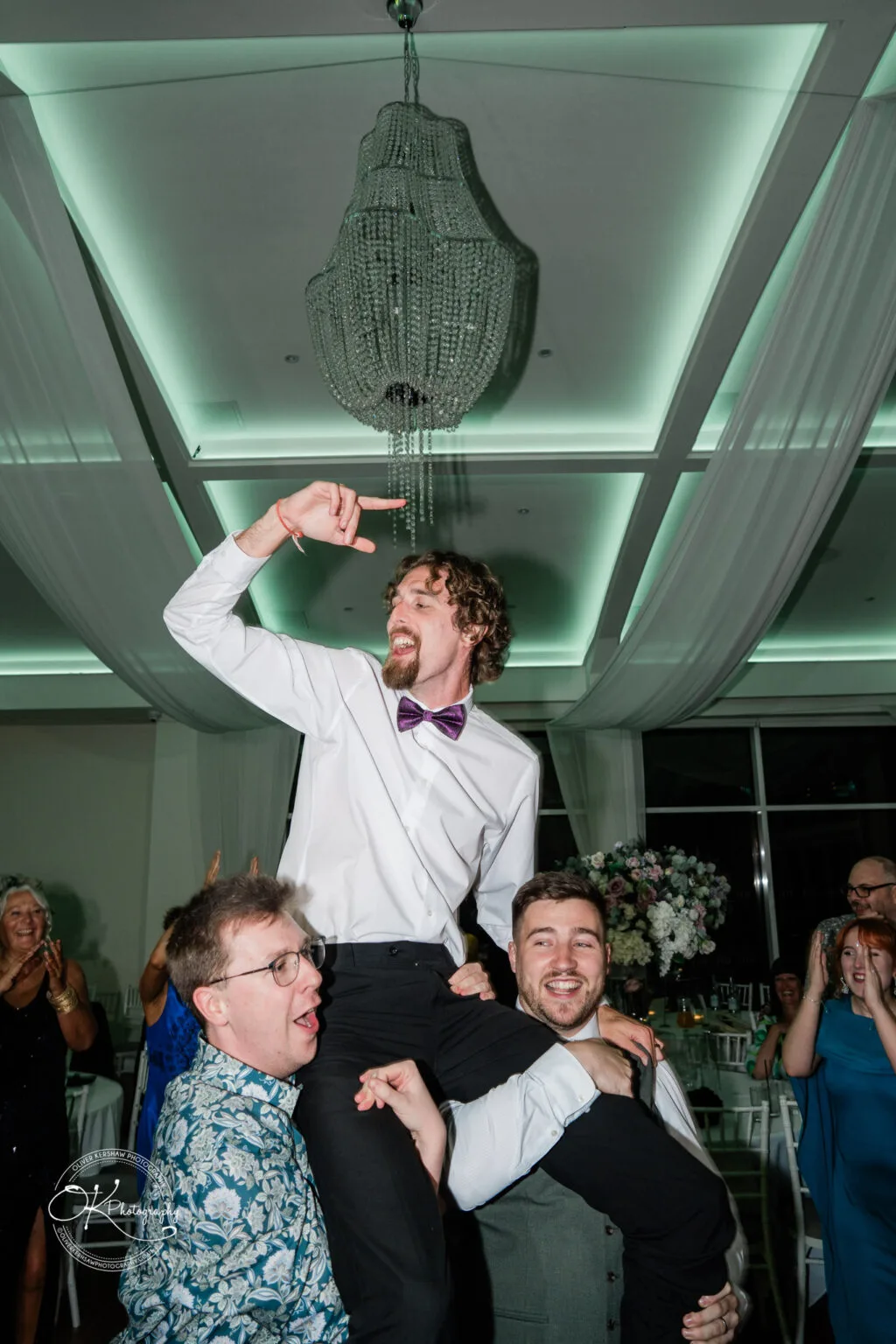 Roshni & Joseph-wedding -Winstanley House-Photography By Oliver Kershaw - OK503479 A groom is being held up by two friends during a wedding celebration at Winstanley House, with guests cheering in the background.