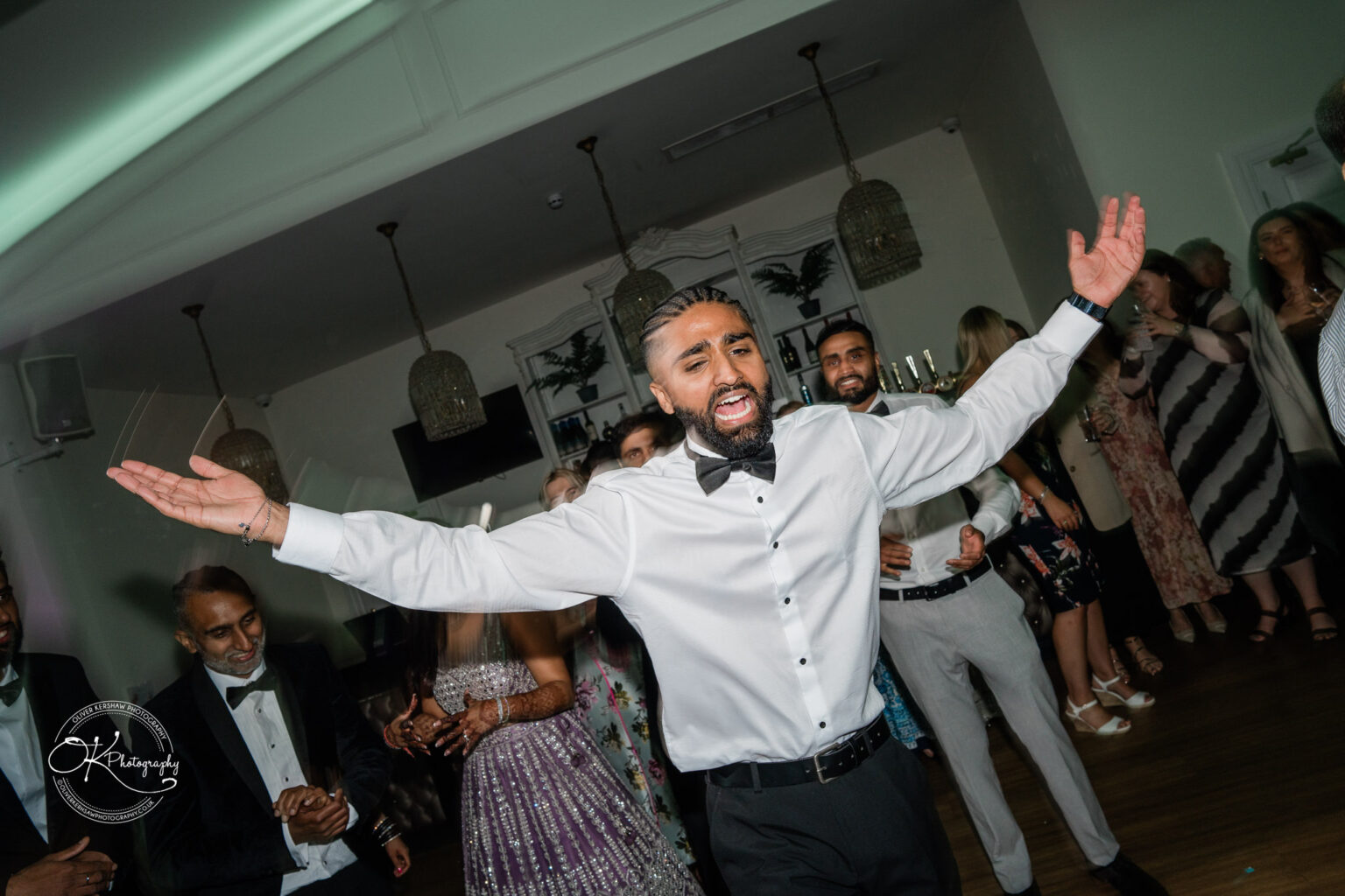 A man in a white shirt and black bow tie joyfully celebrates at a wedding reception, surrounded by guests dancing and enjoying the moment.