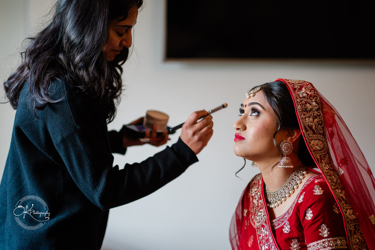 A bride in a red and gold outfit sits patiently as a makeup artist applies finishing touches to her makeup.