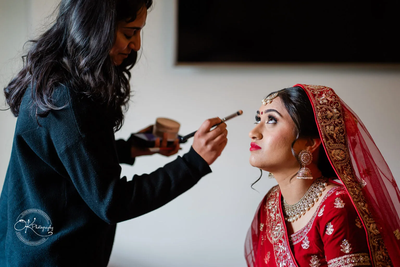 Roshni & Joseph-wedding -Winstanley House-Photography By Oliver Kershaw - OK903971 A bride in a red and gold outfit sits patiently as a makeup artist applies finishing touches to her makeup.