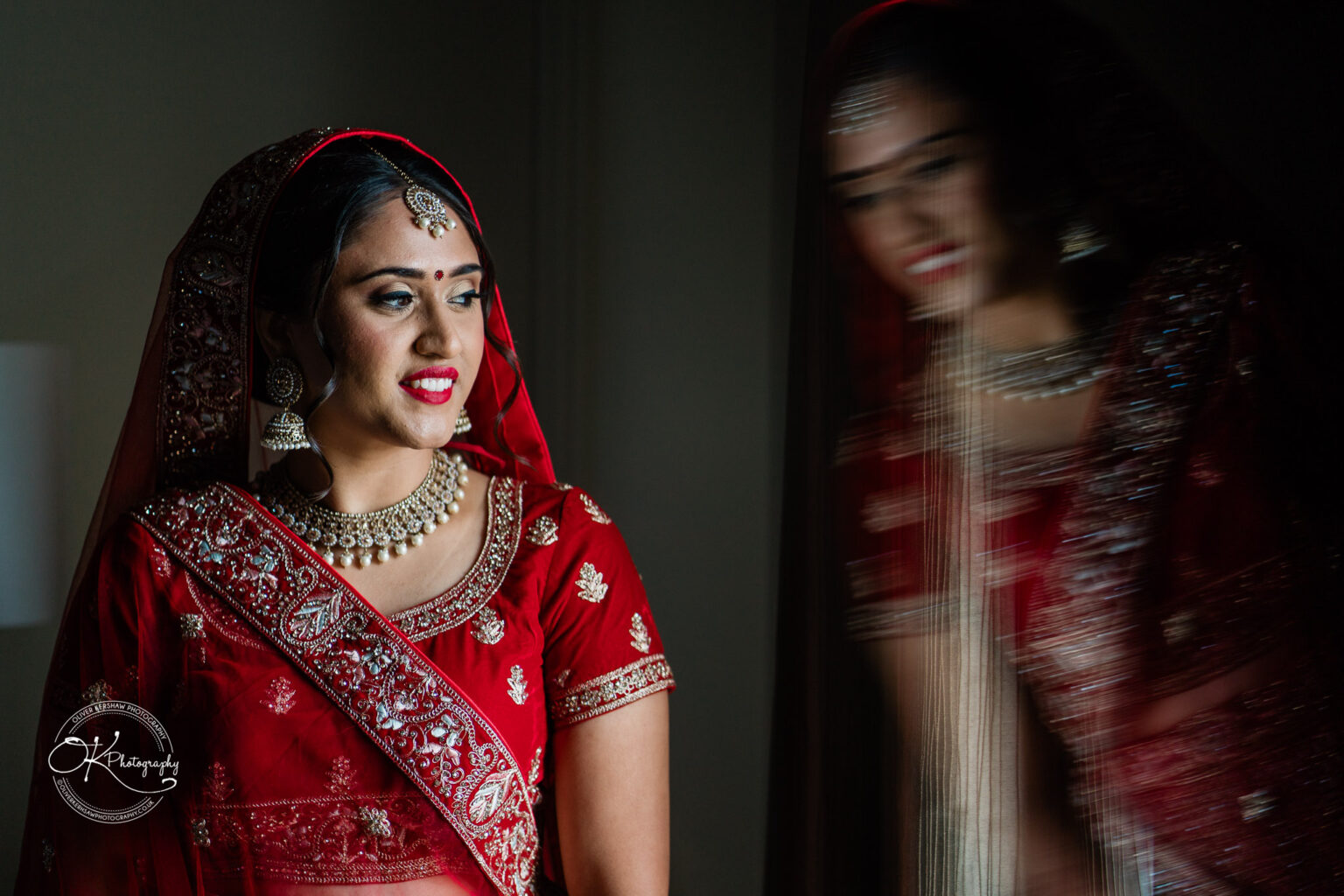 A bride in a red traditional bridal outfit with intricate embroidery and jewelry, smiling softly with a blurred reflection beside her.
