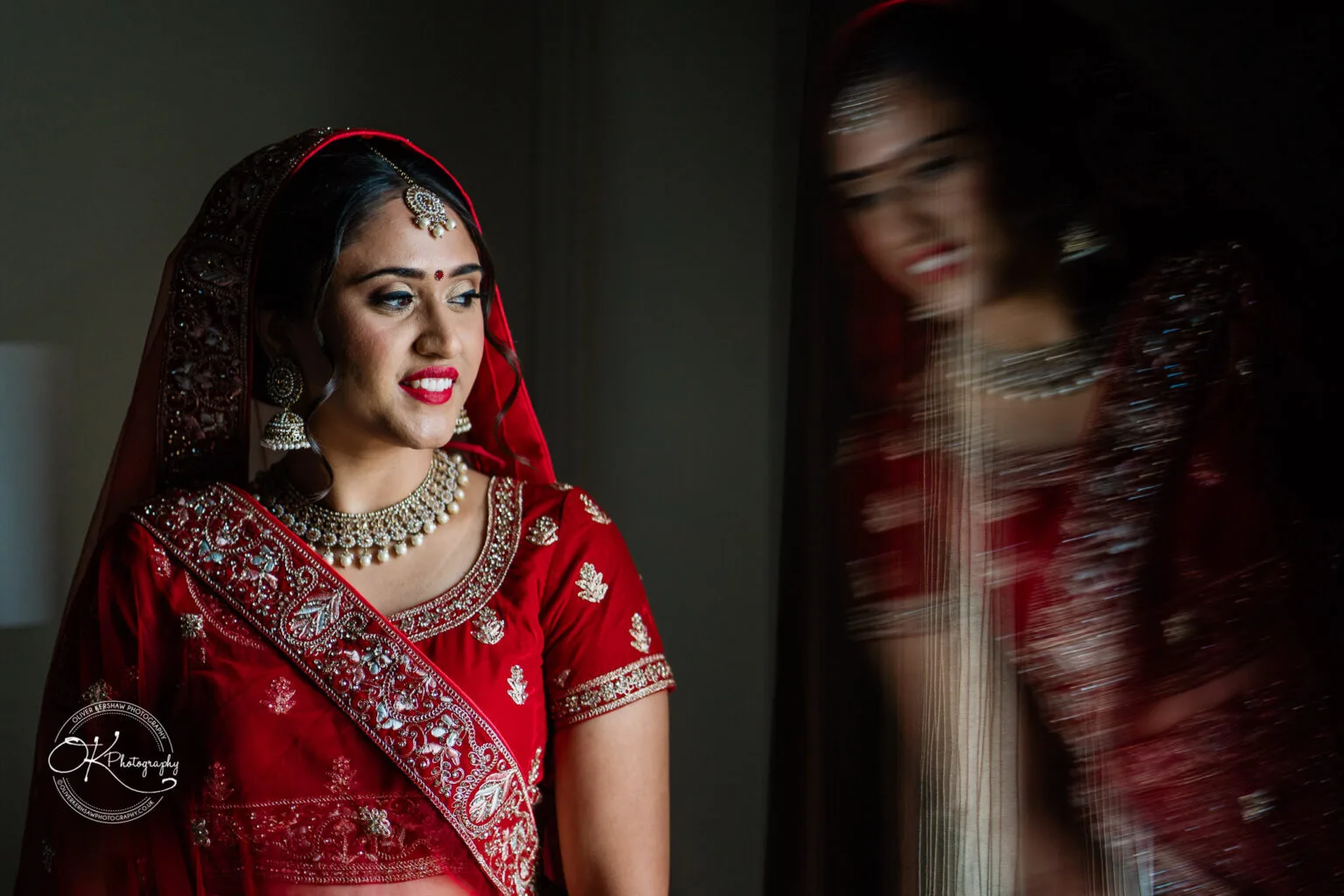 Roshni & Joseph-wedding -Winstanley House-Photography By Oliver Kershaw - OK903992 A bride in a red traditional bridal outfit with intricate embroidery and jewelry, smiling softly with a blurred reflection beside her.