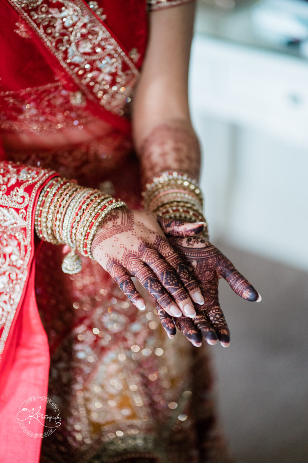 A bride's hands adorned with intricate henna designs and sparkling bangles, showcasing a vibrant red and gold wedding attire.