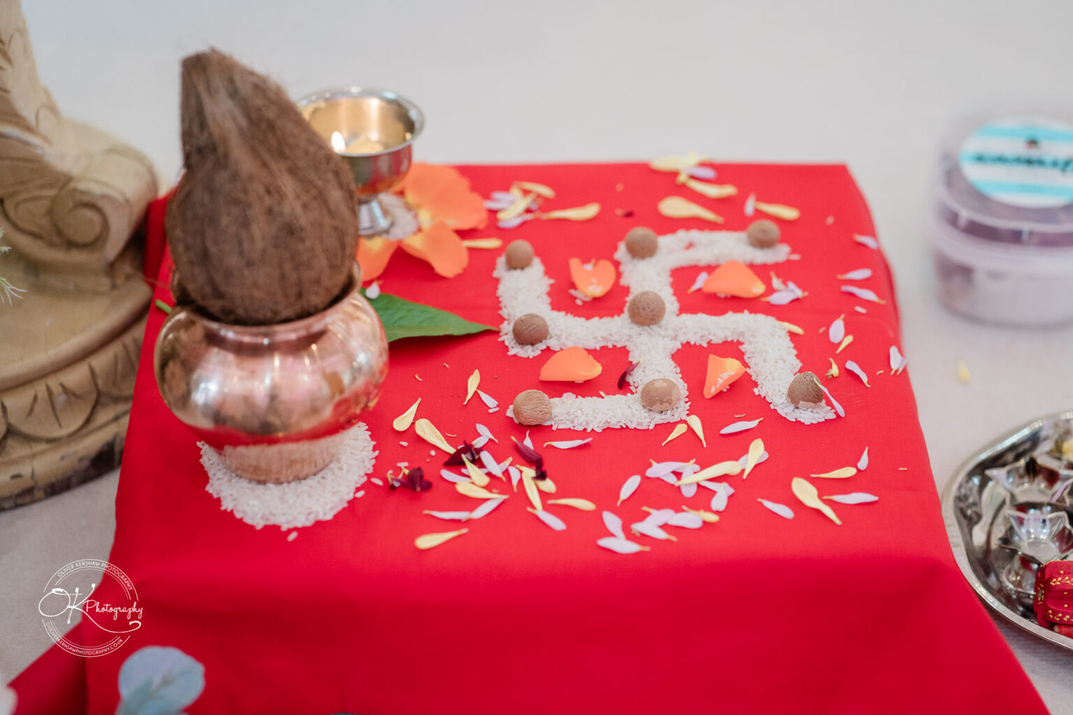 A decorated table featuring a copper pot, coconut, and intricate floral arrangements, with rice and petals forming patterns.