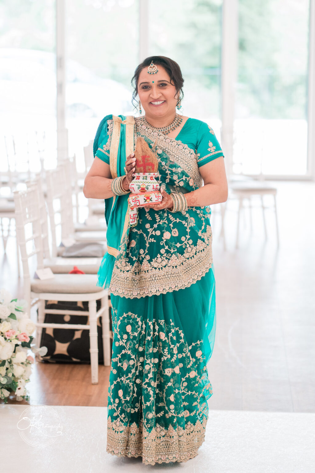 A woman in a turquoise traditional Indian outfit holds a decorative item while smiling, standing in a wedding venue with empty chairs in the background.