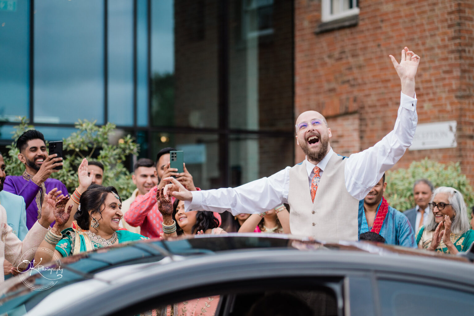 A joyful groom in a vest raises his arms in celebration outside Winstanley House, surrounded by smiling friends and family.