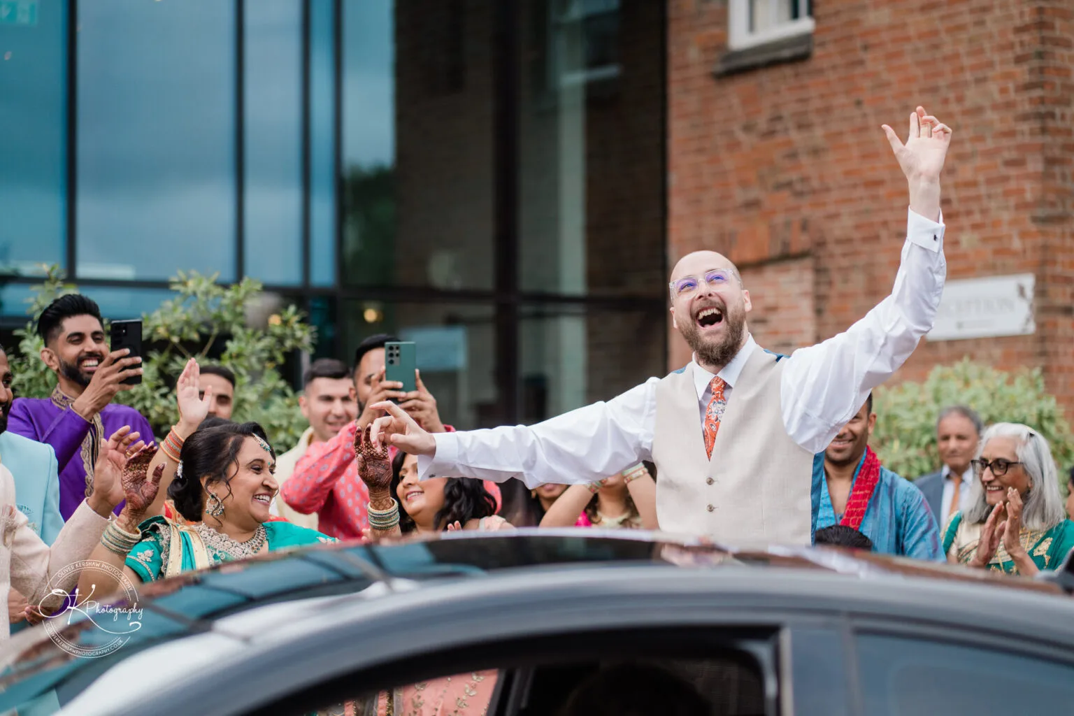 Roshni & Joseph-wedding -Winstanley House-Photography By Oliver Kershaw - OK904114 A joyful groom in a vest raises his arms in celebration outside Winstanley House, surrounded by smiling friends and family.