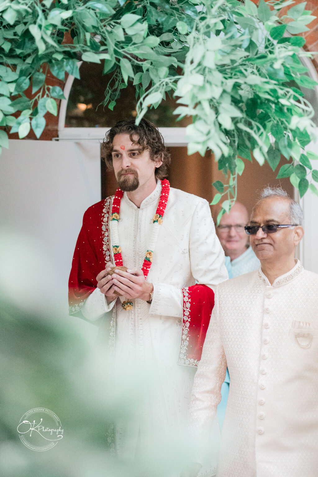 A groom in a traditional white and red wedding outfit stands elegantly with floral garlands, while an elder in formal attire looks on.